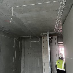 Roman Savchuk - Wnętrze w stanie surowym z widoczną instalacją elektryczną na suficie i ścianach. Pracownik w kasku i kamizelce odblaskowej w tle.