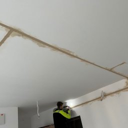 Roman Savchuk - Elektryk w kamizelce montuje instalację elektryczną w bruzdach na suficie, przygotowaną do zalania. Widoczne przewody i rozdzielnica.