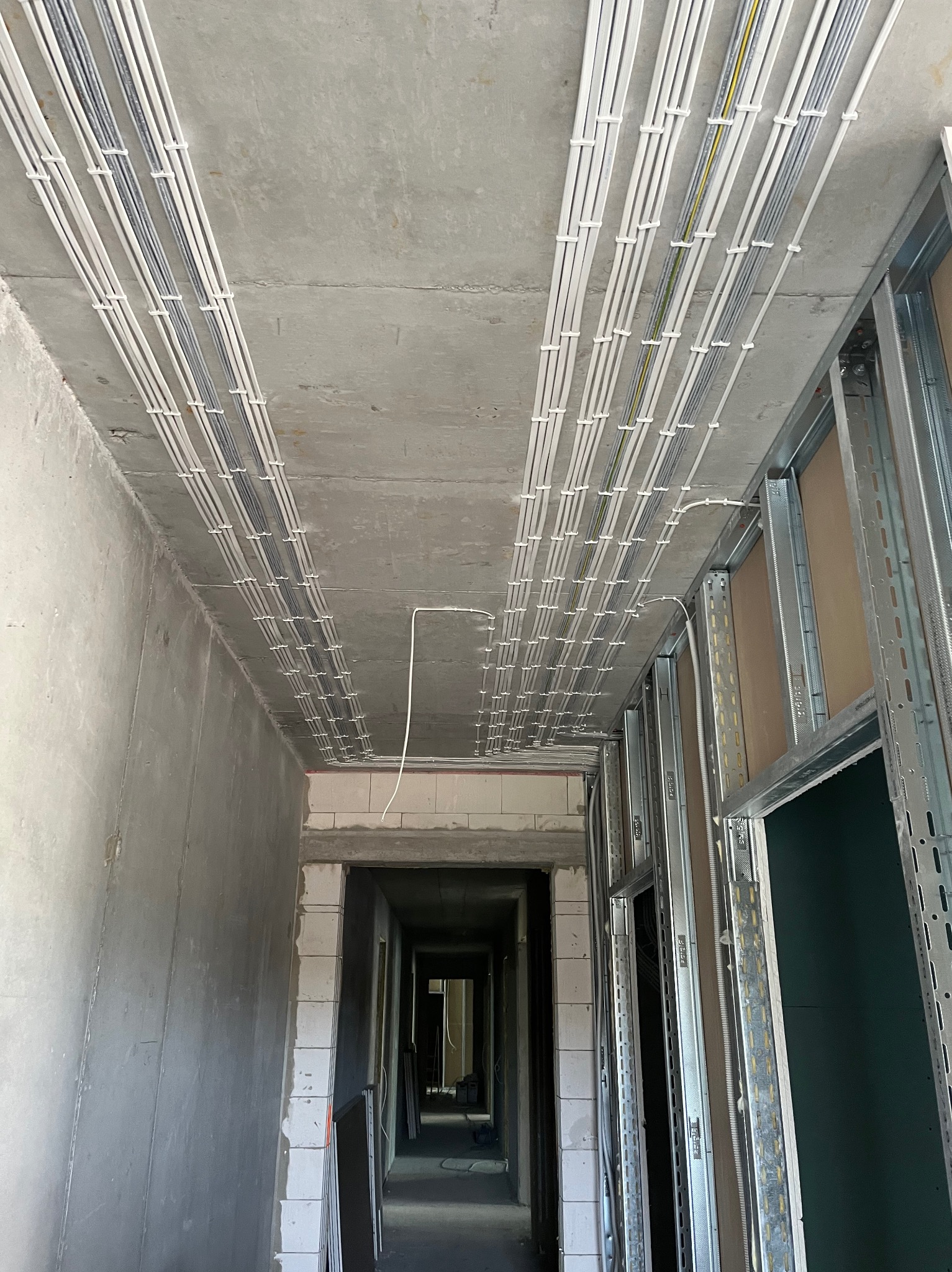 Równolegle ułożone kable w białych rurkach przymocowane do sufitu w surowym wnętrzu budynku w trakcie prac wykończeniowych.