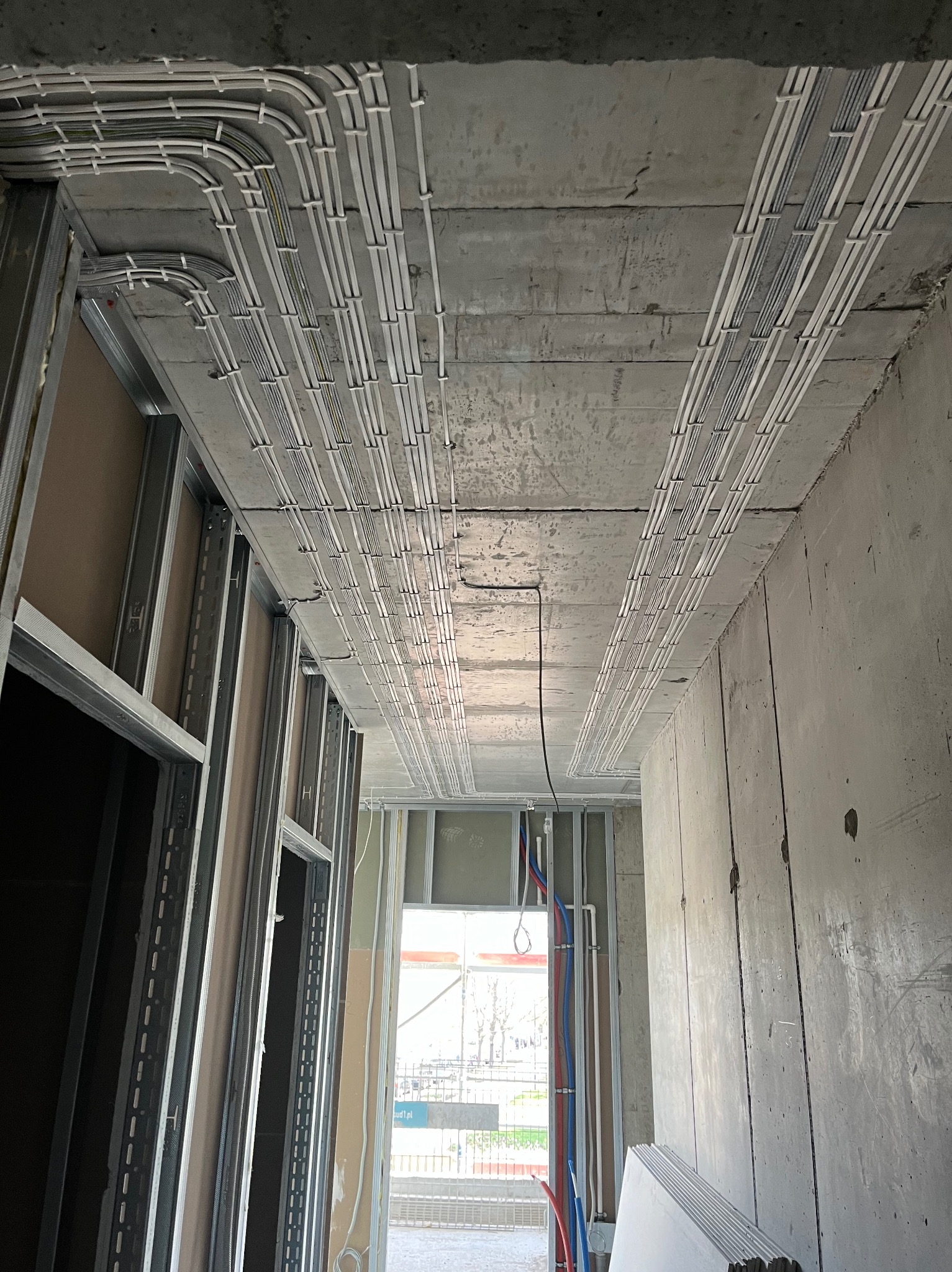 Surowy strop z równolegle ułożonymi kablami w białych rurkach, przytwierdzonymi do betonu. Widoczne metalowe profile ścian działowych i rury instalacji wodnej.