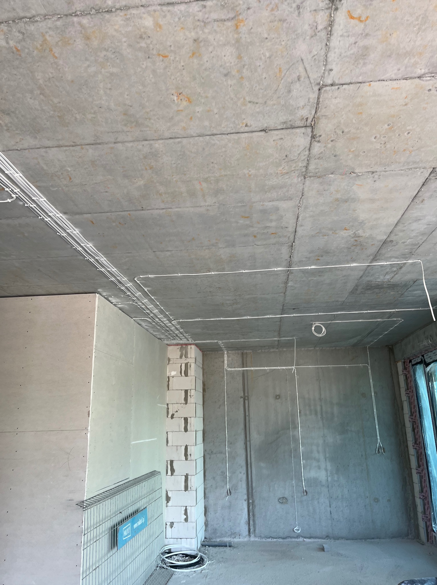 Surowy stan wnętrza z widoczną instalacją elektryczną pod sufitem. Kable prowadzone wzdłuż betonowego sufitu i ścian z pustaków.