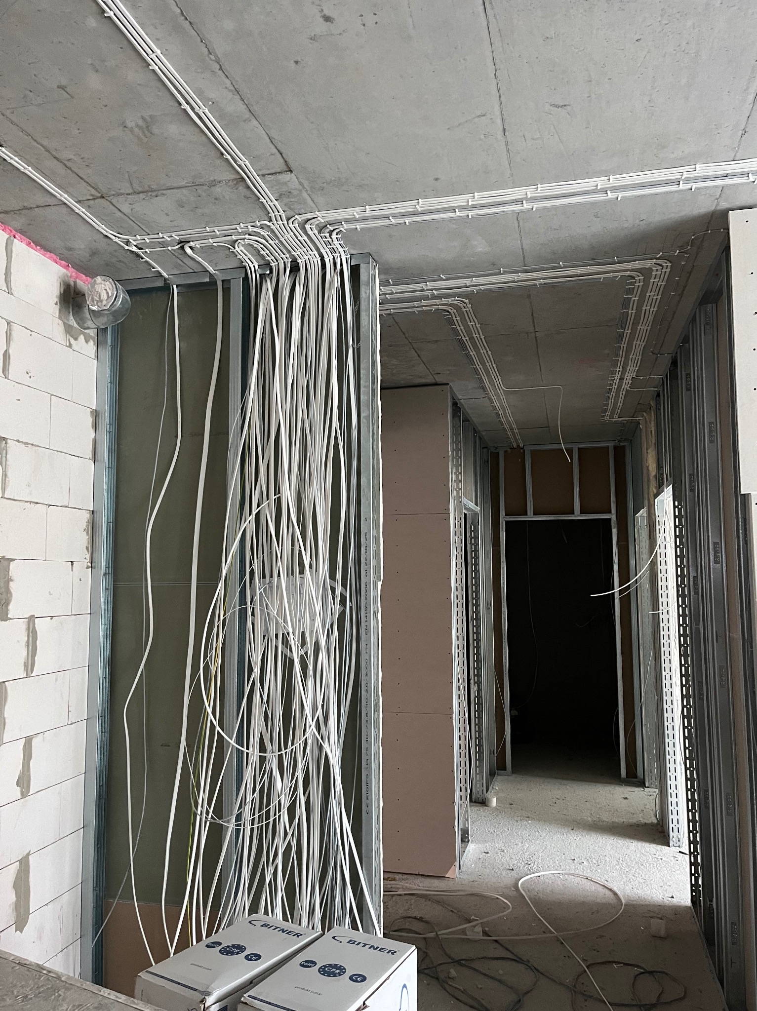 Rozprowadzenie przewodów elektrycznych w nowym budynku, widoczne kable zwisające ze stropu oraz puszki instalacyjne Bitner. Stan surowy, ściany z bloków i profile.