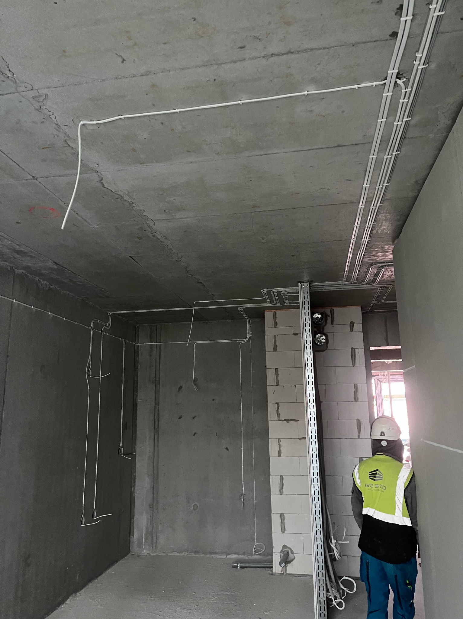 Wnętrze w stanie surowym z widoczną instalacją elektryczną na suficie i ścianach. Pracownik w kasku i kamizelce odblaskowej w tle.