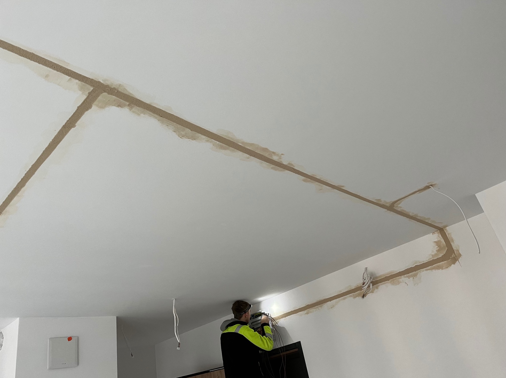 Elektryk w kamizelce montuje instalację elektryczną w bruzdach na suficie, przygotowaną do zalania. Widoczne przewody i rozdzielnica.