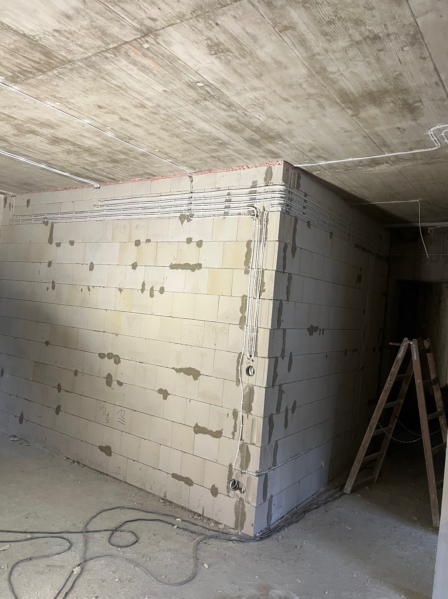 Surowy stan: białe rurki instalacji elektrycznej prowadzone po ścianach z betonu komórkowego w nowym budynku, widoczne kable na podłodze i drabina.