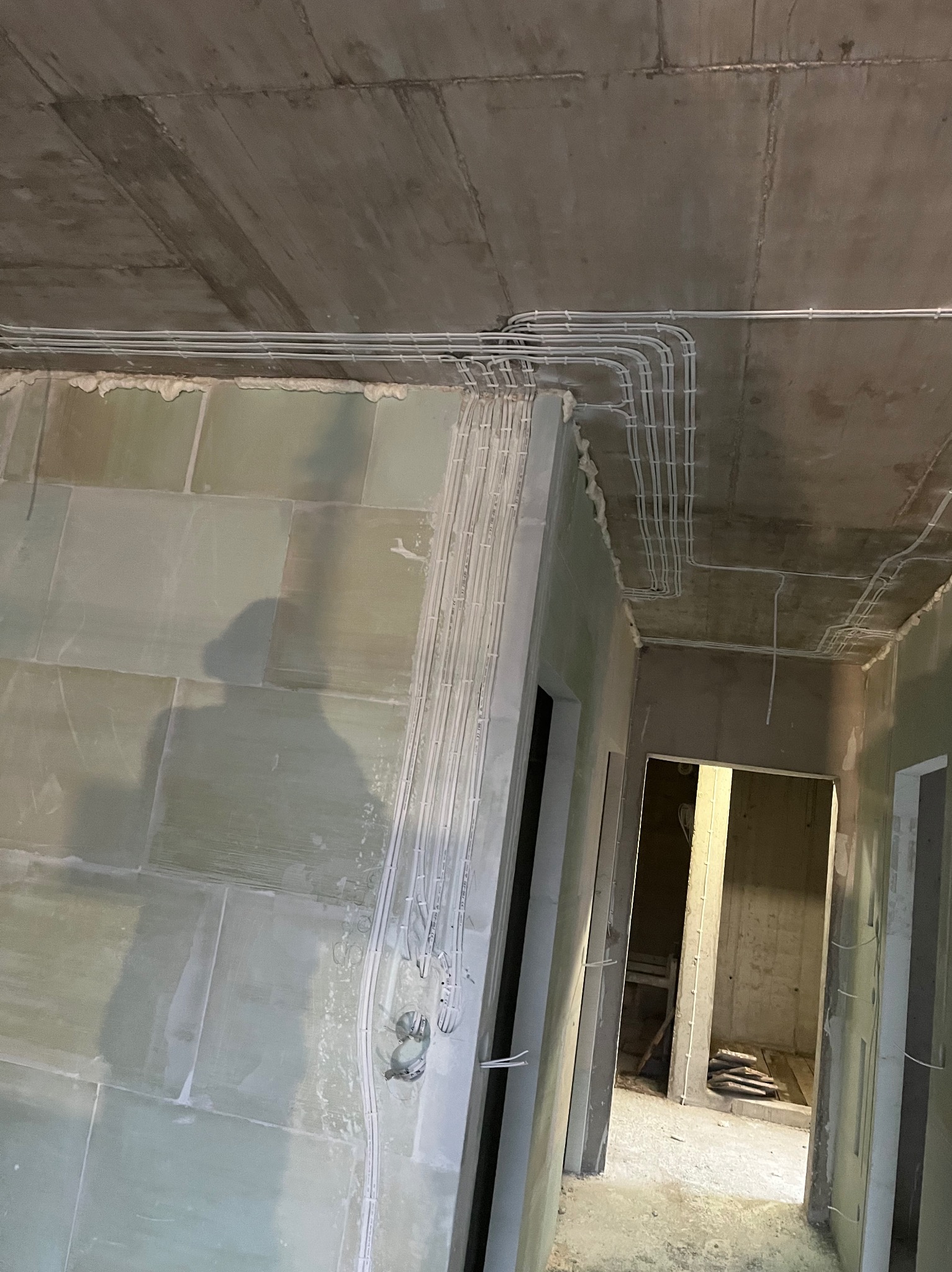 Wnętrze w trakcie prac wykończeniowych: widoczne przewody elektryczne ułożone pod sufitem i wzdłuż ścian, zarysowane otwory drzwiowe, surowy beton sufitu i zielone płyty gipsowe.