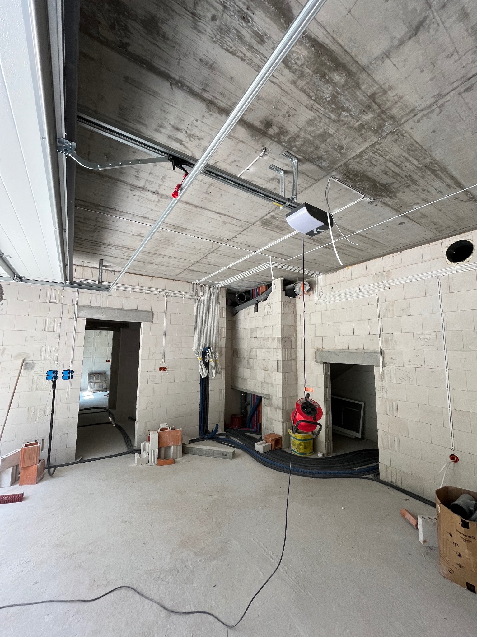 Surowy stan wnętrza: widoczne instalacje elektryczne w białych rurkach, otwory drzwiowe w ścianach z betonu komórkowego, sufit betonowy z zamontowanym napędem do bramy garażowej.