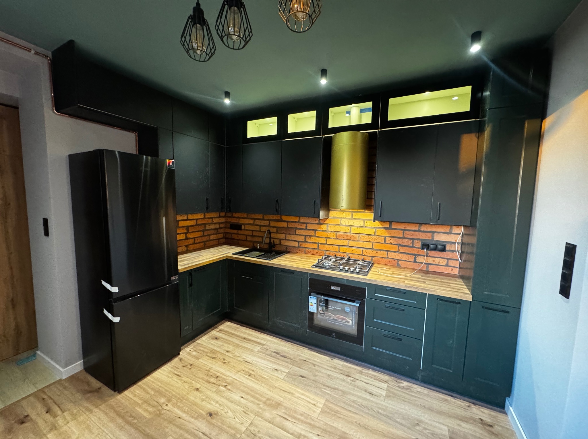 Nowoczesna kuchnia w odcieniach zieleni z ceglaną ścianą, drewnianym blatem i czarnym sprzętem AGD. Widoczne designerskie lampy wiszące i oświetlenie podszafkowe.