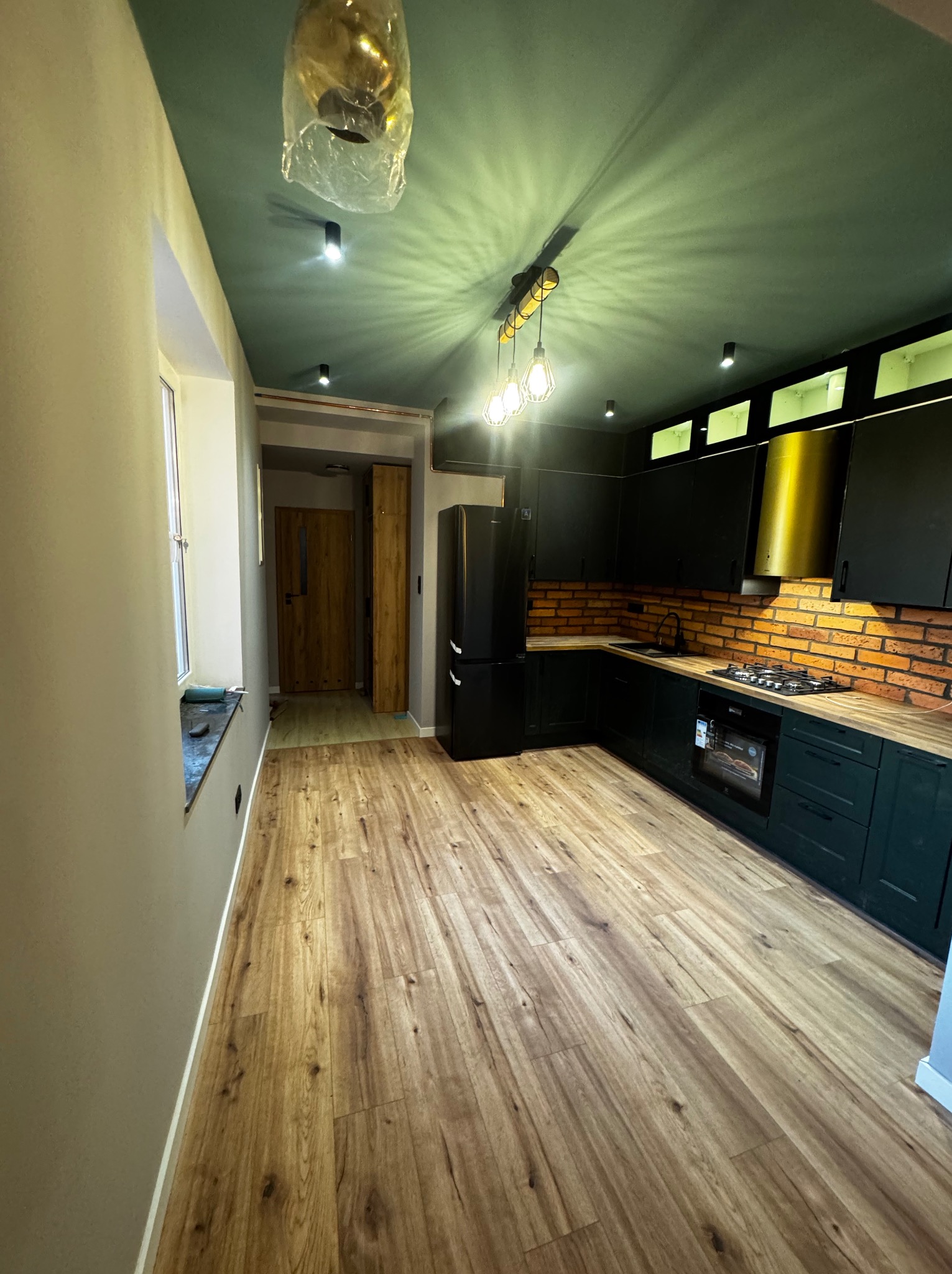 Nowoczesna, wykończona kuchnia z ciemnozielonymi szafkami, drewnianym blatem i ceglaną ścianą. Widoczny czarny sprzęt AGD i drewniana podłoga. Sufit w odcieniach zieleni.