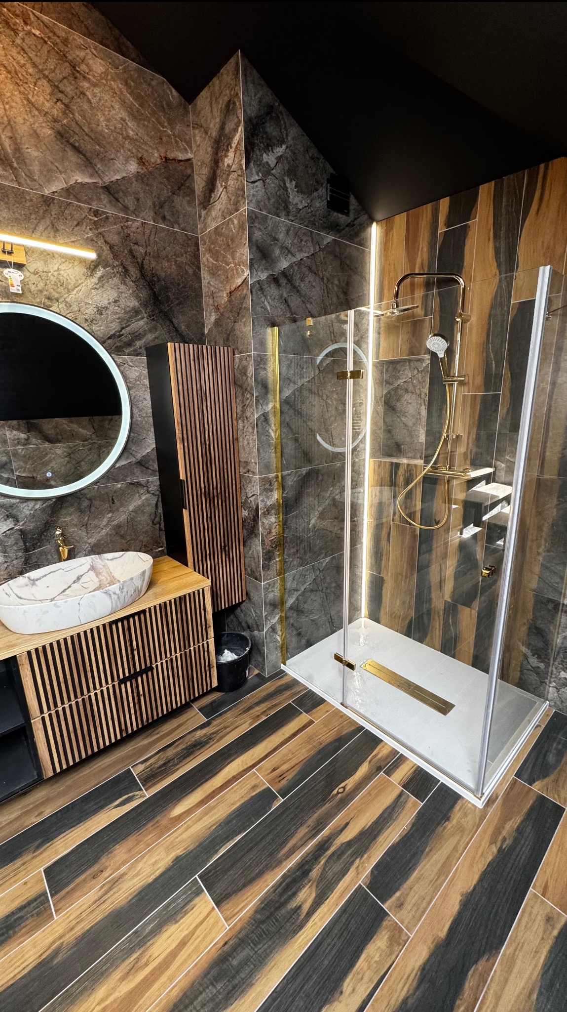 Nowoczesna łazienka z kabiną prysznicową, marmurową umywalką i drewnianymi akcentami. Ciemne płytki imitujące kamień na ścianach i podłodze.
