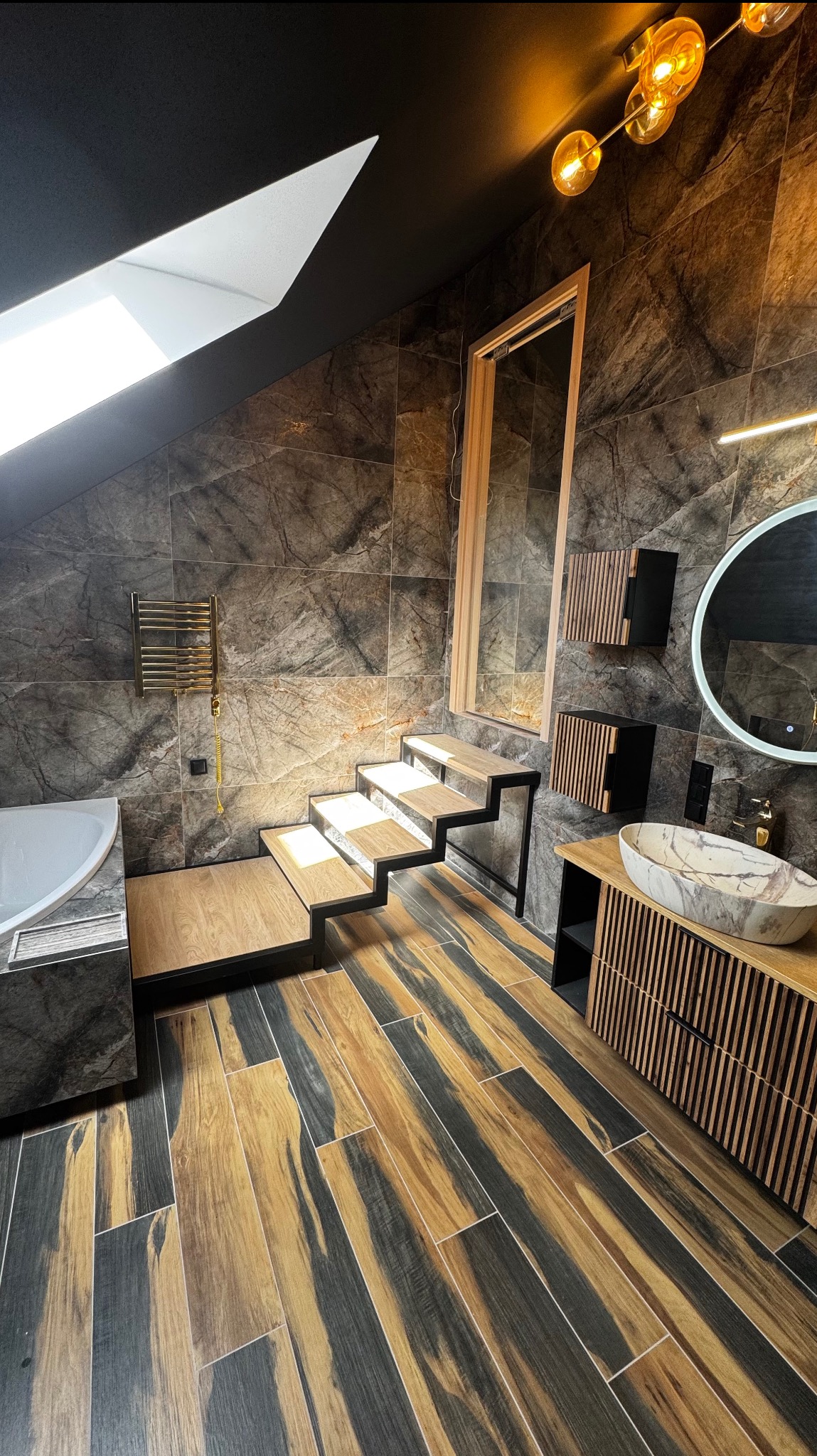Stylowa łazienka na poddaszu z marmurowymi płytkami, drewnianą podłogą i schodami. Nowoczesna umywalka i okrągłe lustro z podświetleniem LED.