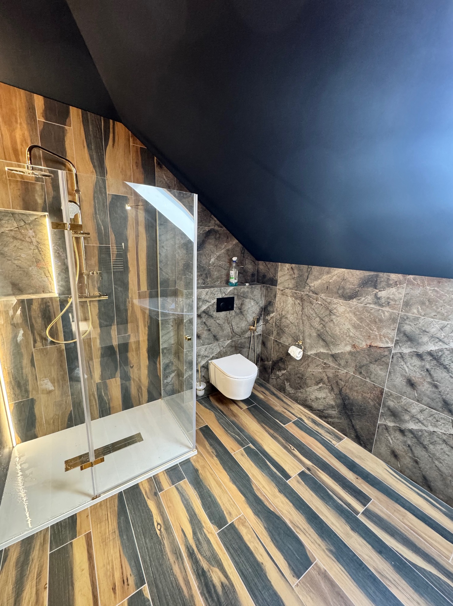 Nowoczesna łazienka na poddaszu z kabiną prysznicową typu walk-in, wykończona płytkami imitującymi marmur i drewno. Czarny sufit. Złote akcenty w armaturze.