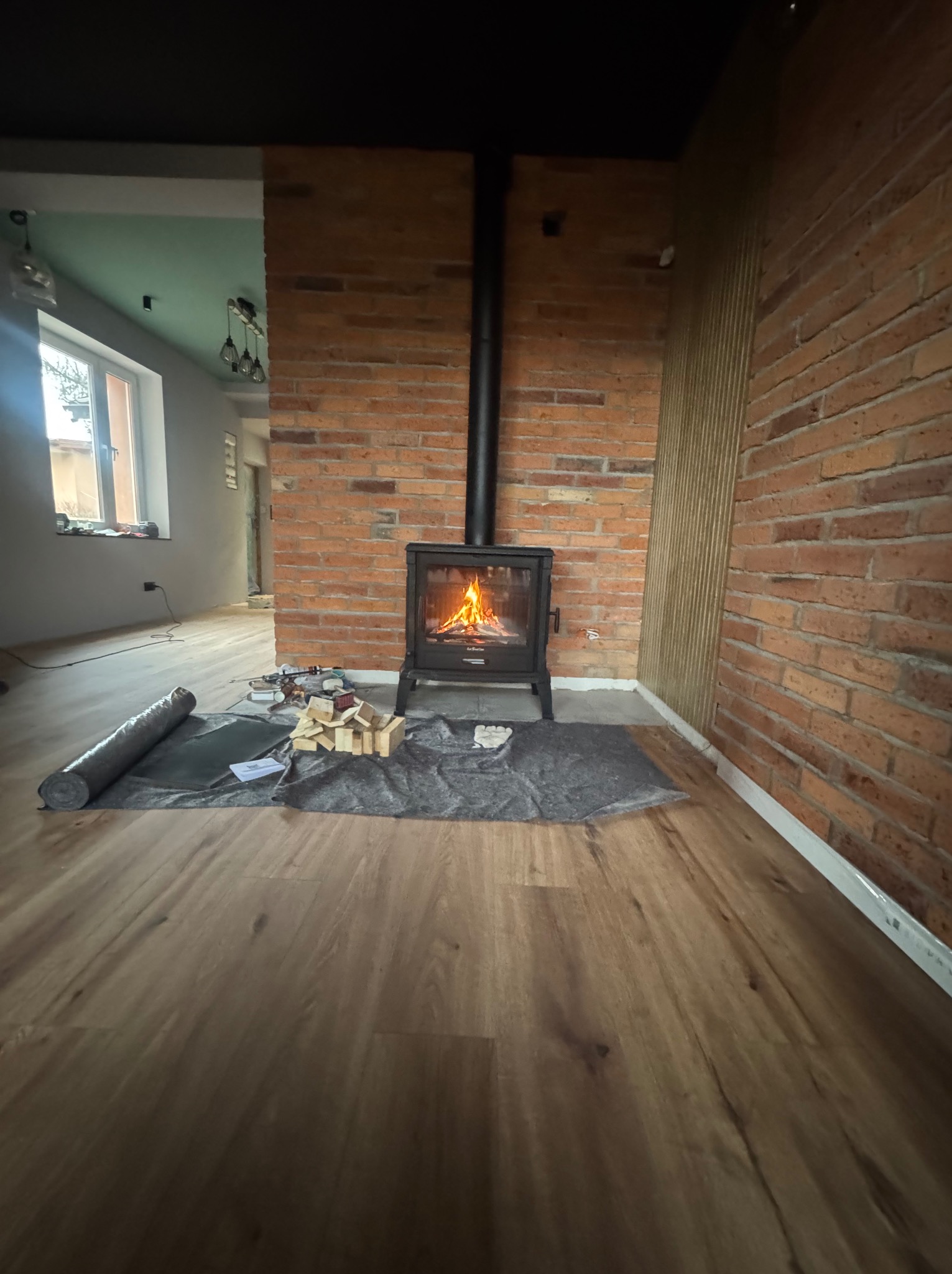 Wnętrze z ceglaną ścianą i kominkiem typu koza. Na podłodze panele, widoczne materiały przygotowane do montażu. Częściowo widoczny inny pokój z oknem.