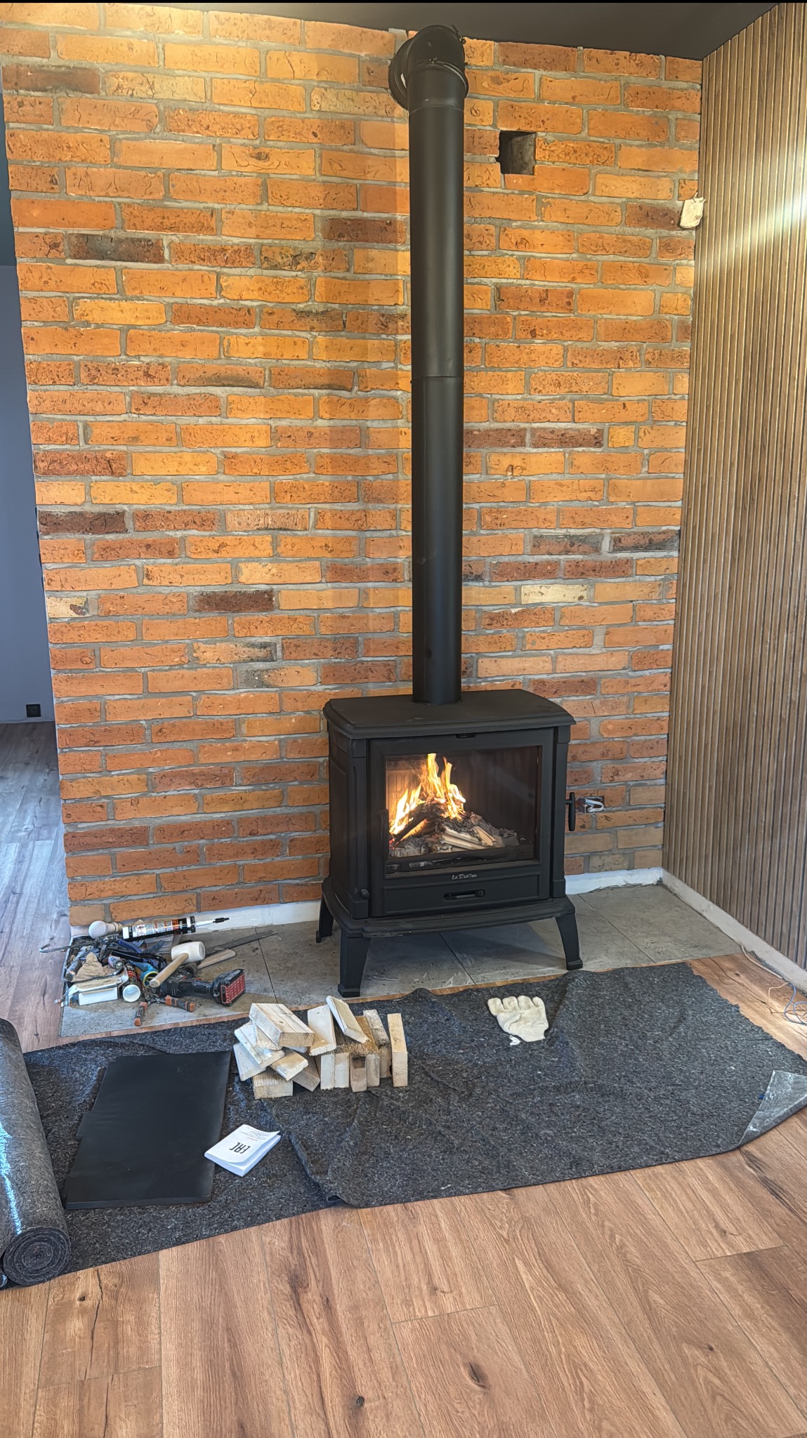 Montaż pieca wolnostojącego z czarnym kominem na tle ściany z cegły. Widoczne narzędzia i materiały przygotowane do instalacji. Płomienie w palenisku.