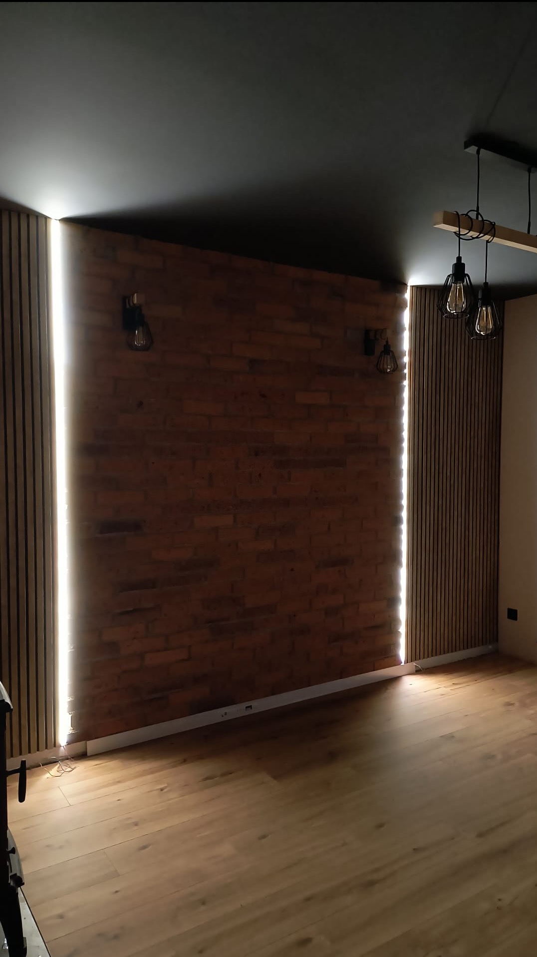 Pokój z ceglaną ścianą, drewnianymi panelami z oświetleniem LED, lampami wiszącymi i drewnianą podłogą. Ciemny sufit dodaje kontrastu.
