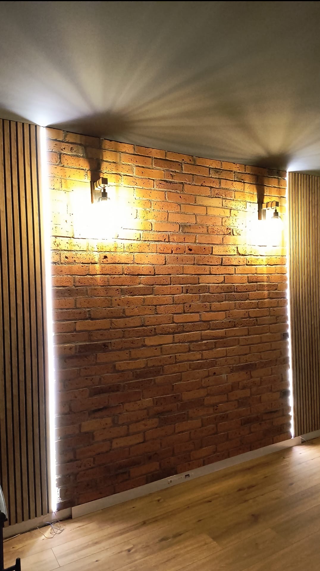 Ceglana ściana z oświetleniem LED po bokach i dwoma lampami na górze. Dekoracyjne panele ścienne po bokach. Parkiet na podłodze. Ciepłe światło.