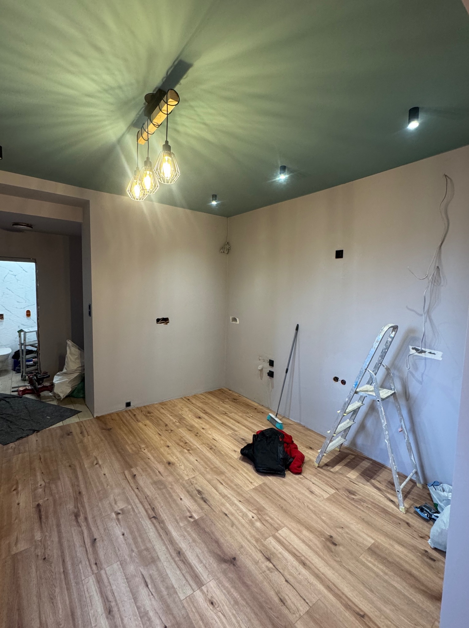 Wnętrze w trakcie prac wykończeniowych: drewnopodobna podłoga, pomalowany sufit na zielono, drabina oparta o ścianę, widoczne przewody elektryczne, nowoczesna lampa.