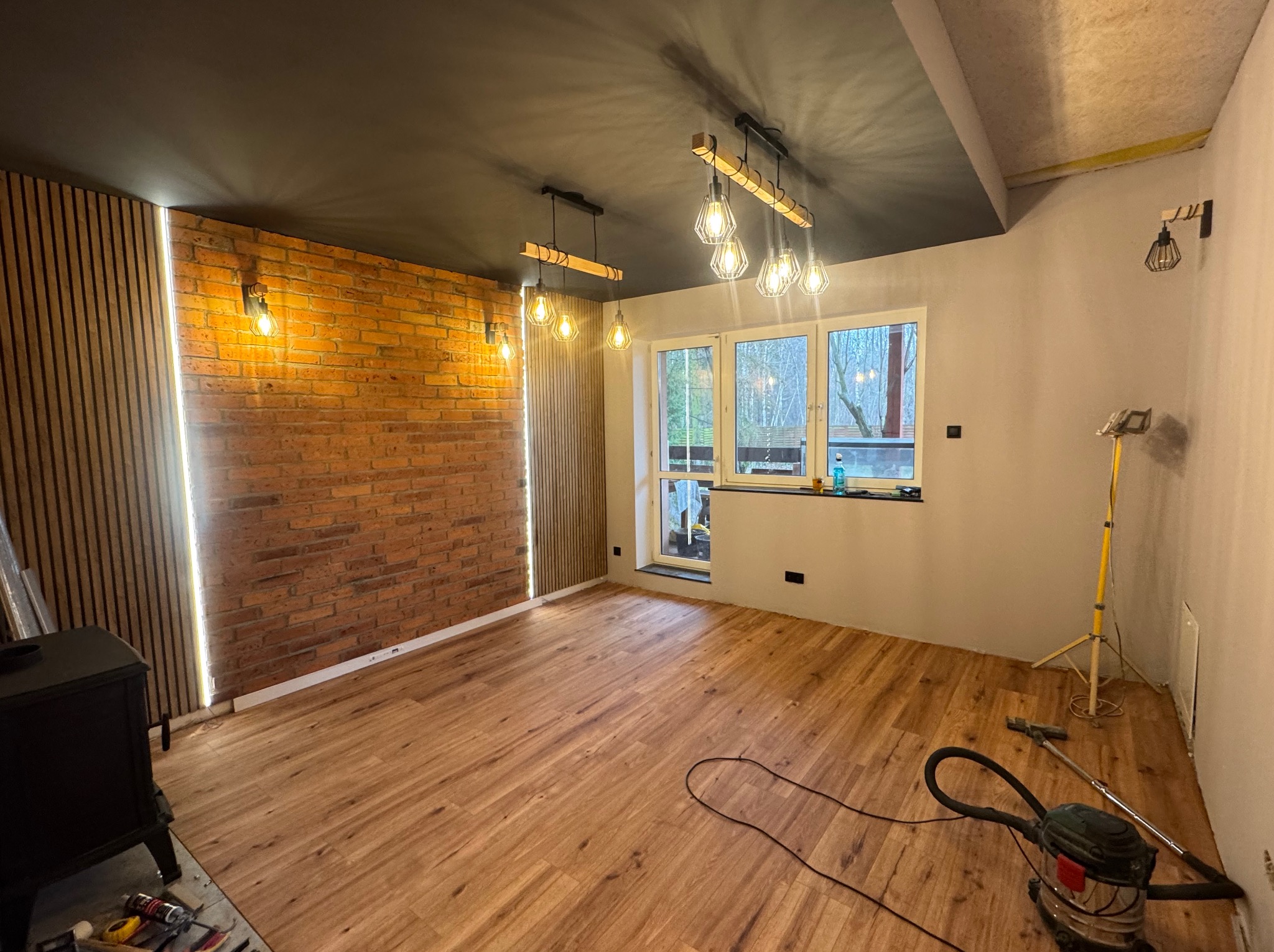 Wykończone wnętrze pokoju z drewnianą podłogą, ceglaną ścianą z oświetleniem LED, czarnym sufitem i nowoczesnymi lampami wiszącymi. Widoczny odkurzacz i narzędzia.