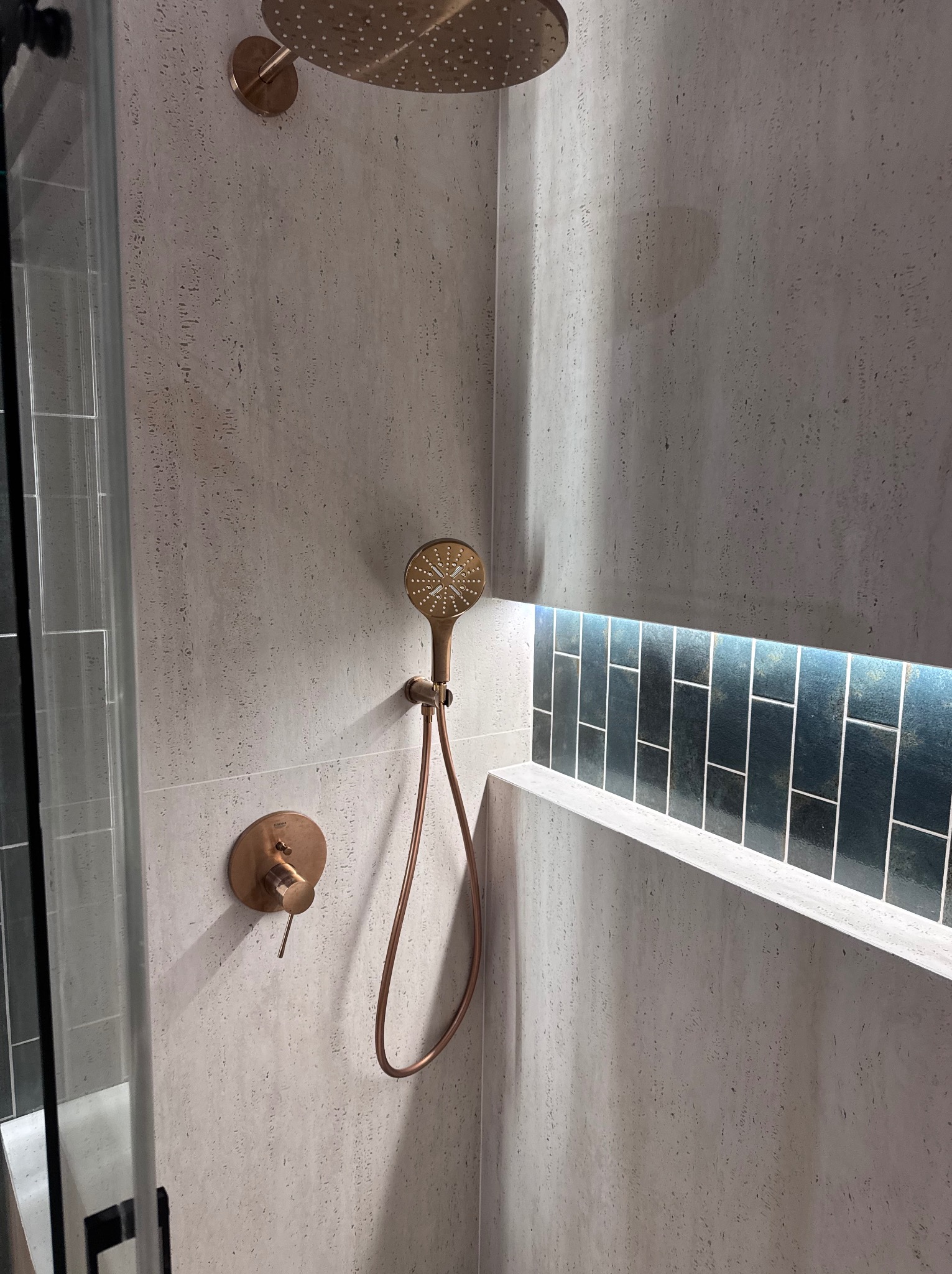 Nowoczesna kabina prysznicowa z armaturą w kolorze miedzi i podświetlaną wnęką z ciemnymi płytkami. Minimalistyczny design, jasne płytki imitujące kamień.