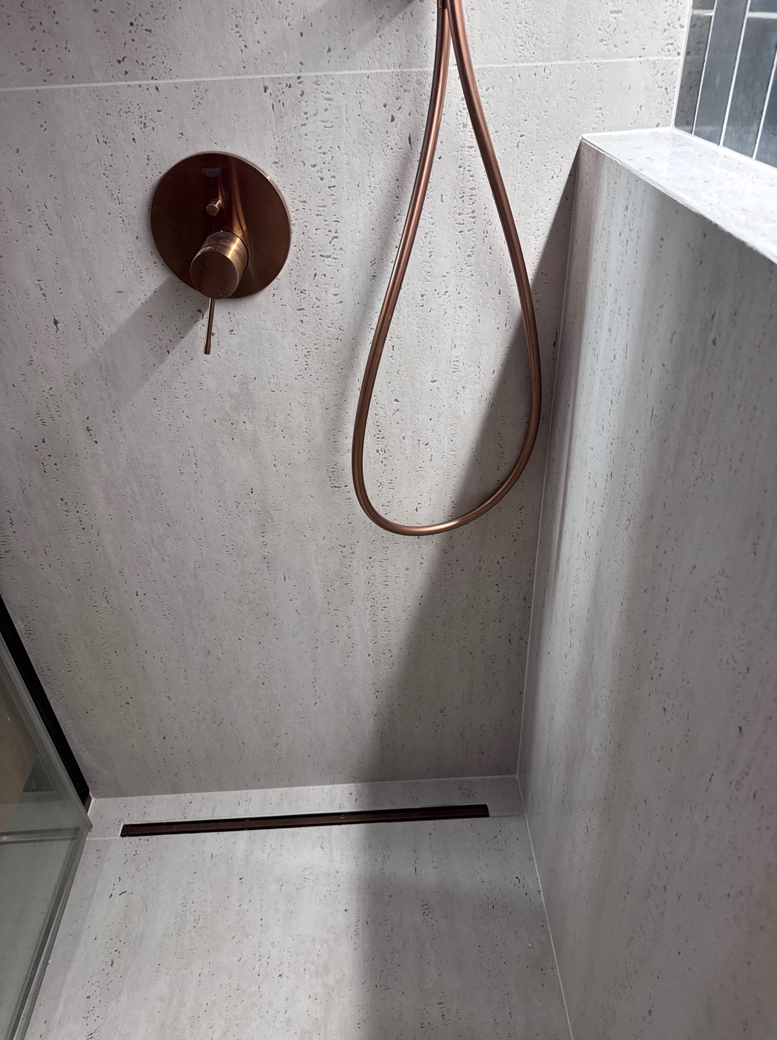 Minimalistyczna kabina prysznicowa z armaturą w kolorze miedzi i szarymi płytkami o porowatej strukturze. Ujęcie z dołu, podkreślające detale wykończenia.