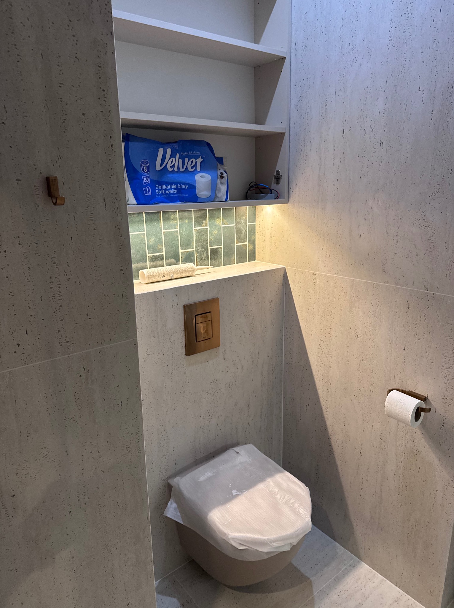 Wykończona toaleta w beżowych płytkach z mozaiką nad spłuczką. Widoczna toaleta z zabezpieczeniem, uchwyt na papier i szafka z zapasem papieru toaletowego Velvet.