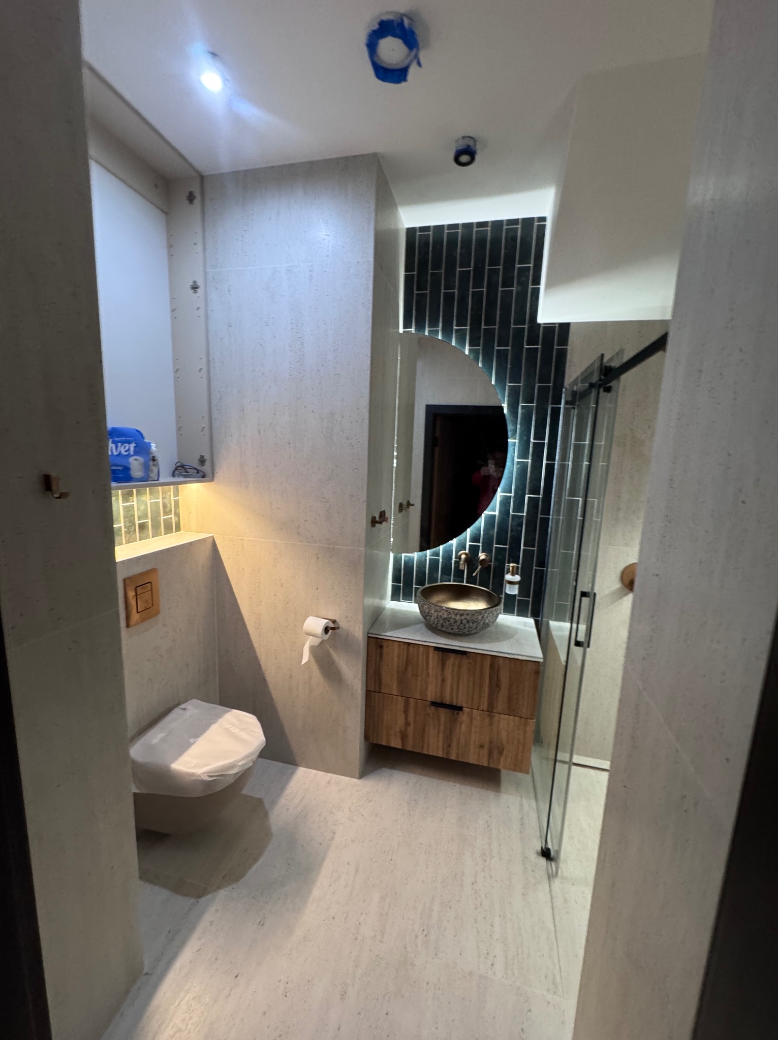 Nowoczesna łazienka z okrągłym lustrem podświetlanym LED, drewnianą szafką pod umywalką i kabiną prysznicową ze szklanymi drzwiami przesuwnymi.