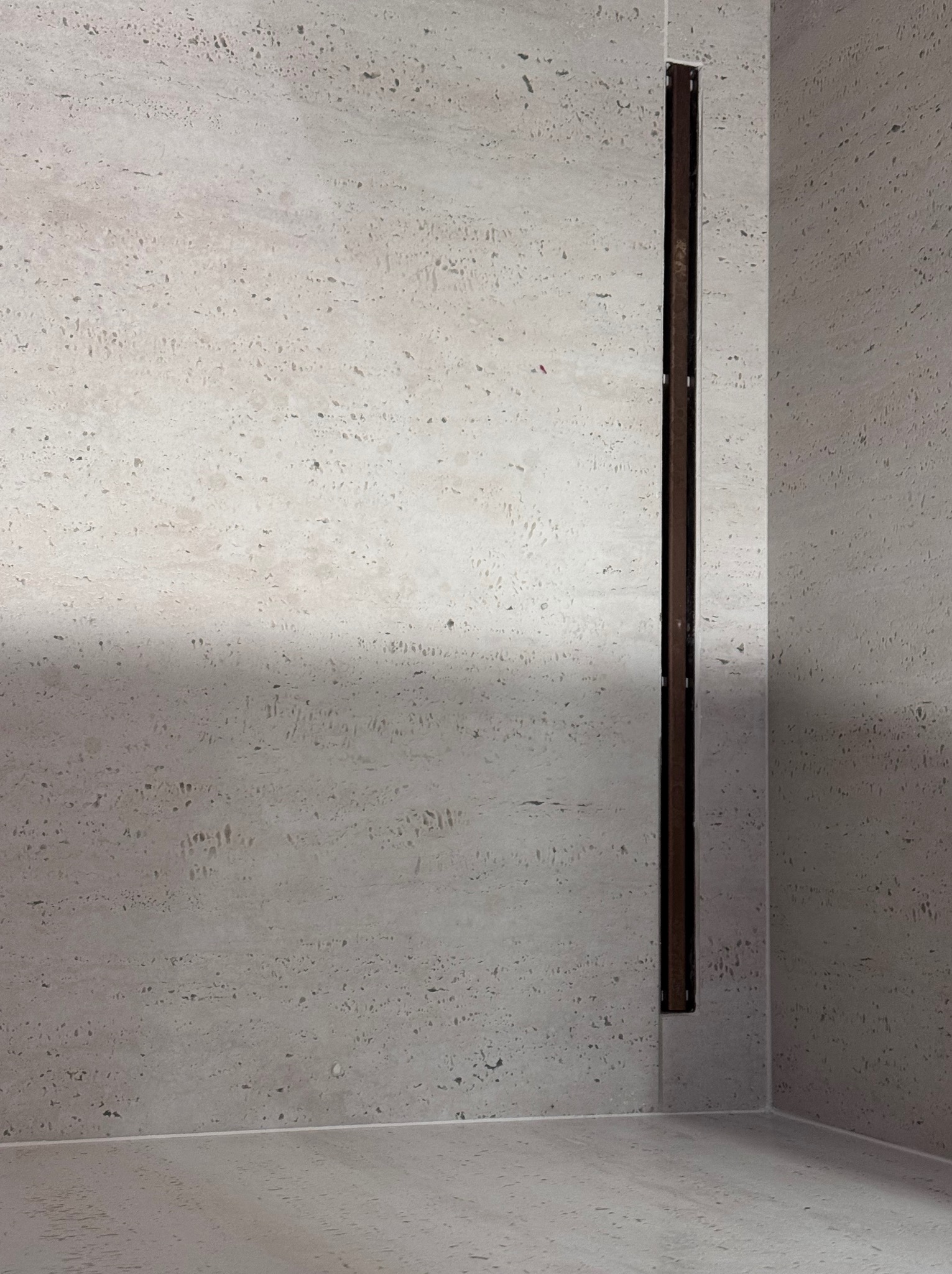 Wykończona ściana i podłoga w łazience z beżowego trawertynu z ukrytym odpływem liniowym. Minimalistyczny design, neutralna kolorystyka.