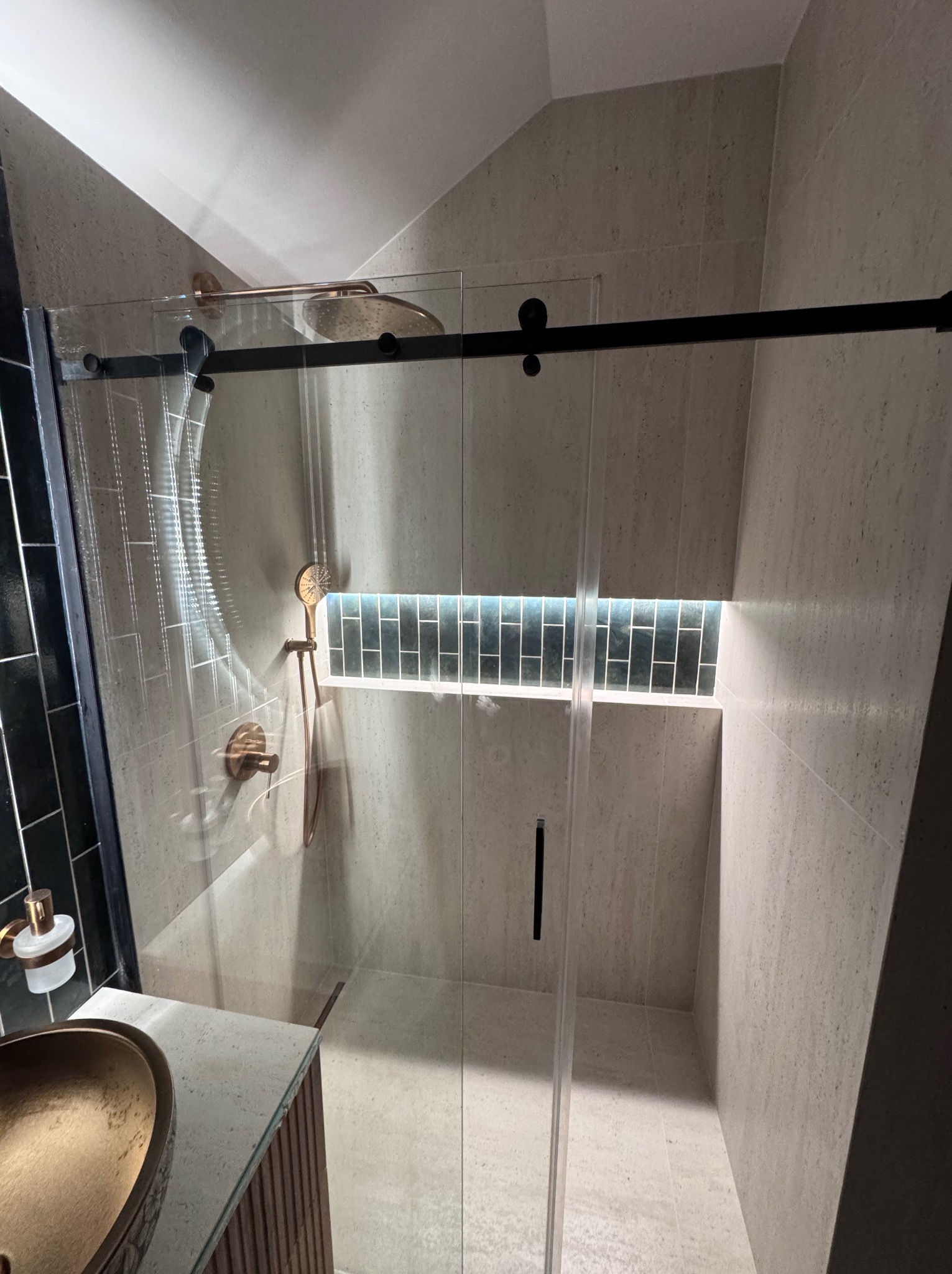 Nowoczesna kabina prysznicowa z przesuwnymi drzwiami, wykończona w beżowych płytkach. Widoczny złoty zestaw prysznicowy i umywalka. Podświetlana wnęka z ciemnymi płytkami.