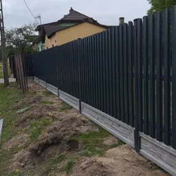 Nowoczesne ogrodzenie metalowe z podmurówką w Różycy. Pionowe panele w kolorze antracytowym, betonowa podmurówka imitująca kamień. Świeżo zamontowane, widoczne ślady prac ziemnych.