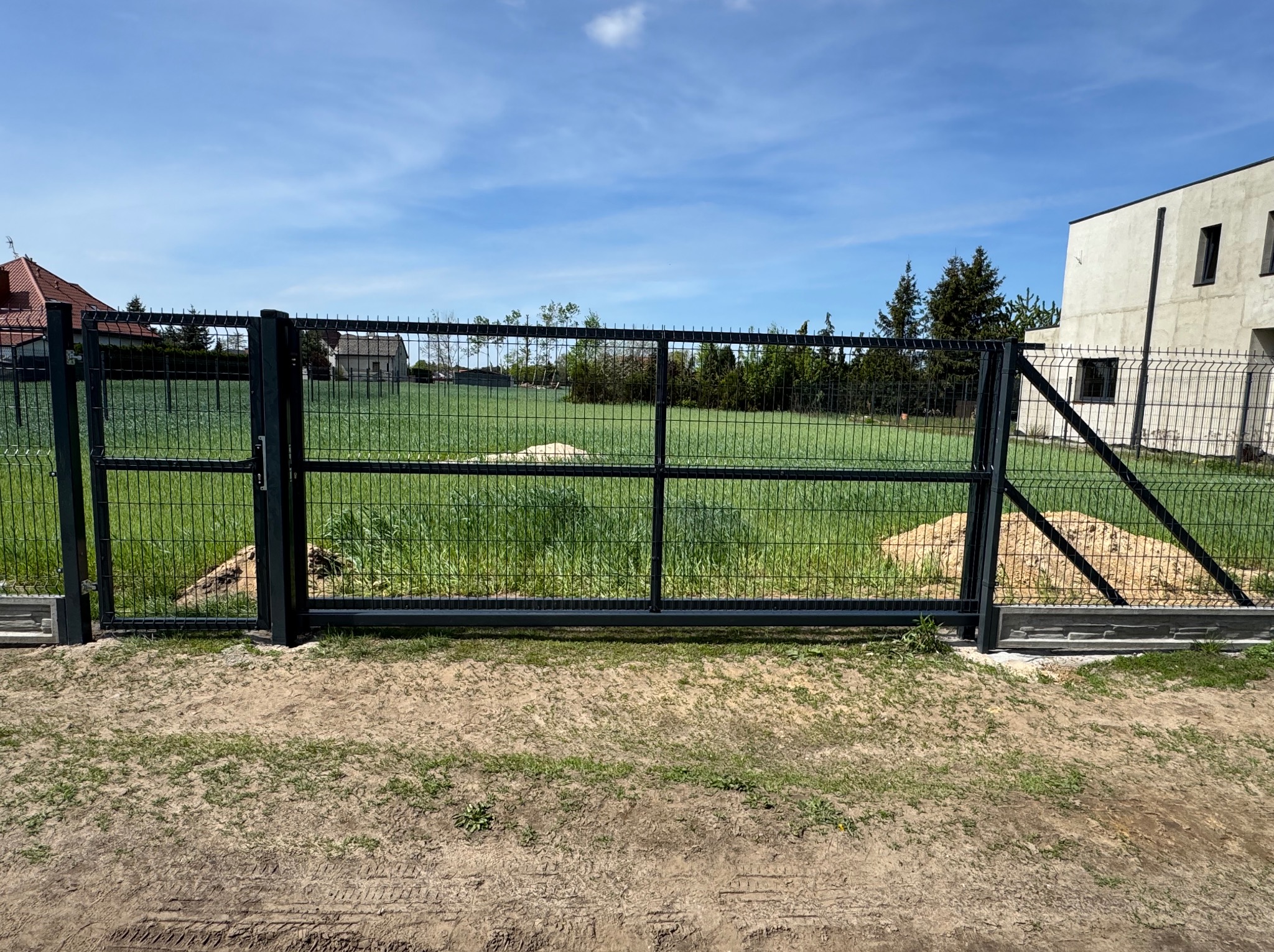 Nowoczesna, grafitowa brama panelowa z widokiem na zielony teren i domy w tle, w słoneczny dzień. Brama zamknięta, z widocznymi elementami betonowymi u podstawy.