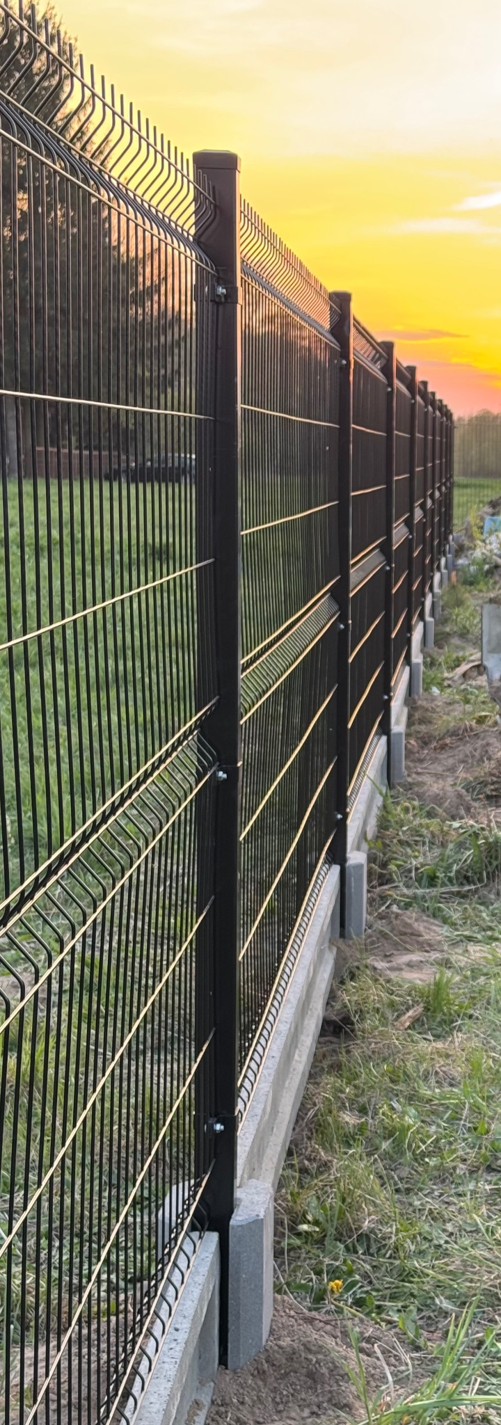 Nowoczesne ogrodzenie panelowe z podmurówką betonową, widziane pod kątem, oświetlone ciepłym światłem zachodzącego słońca. Widoczne mocowania paneli do słupków.