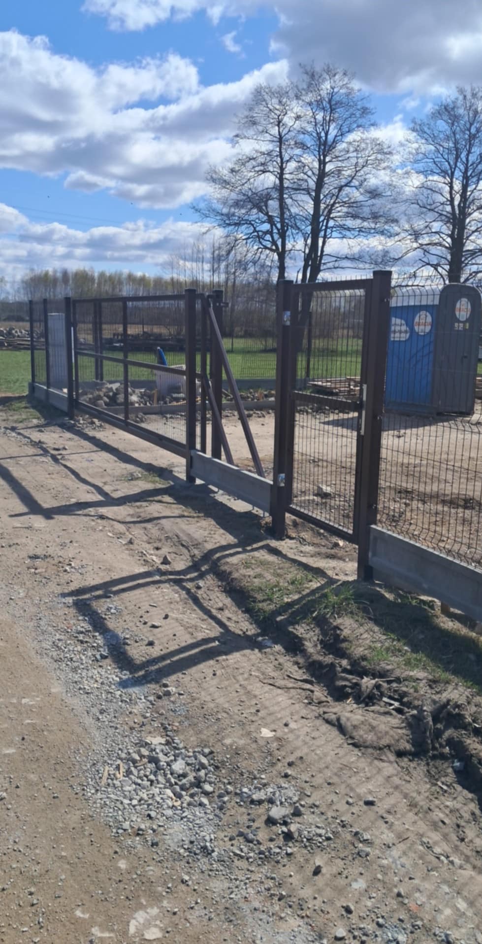 Ciemny panelowy płot z otwartą bramą, betonowa podmurówka, teren budowy, błękitne niebo z chmurami w tle.