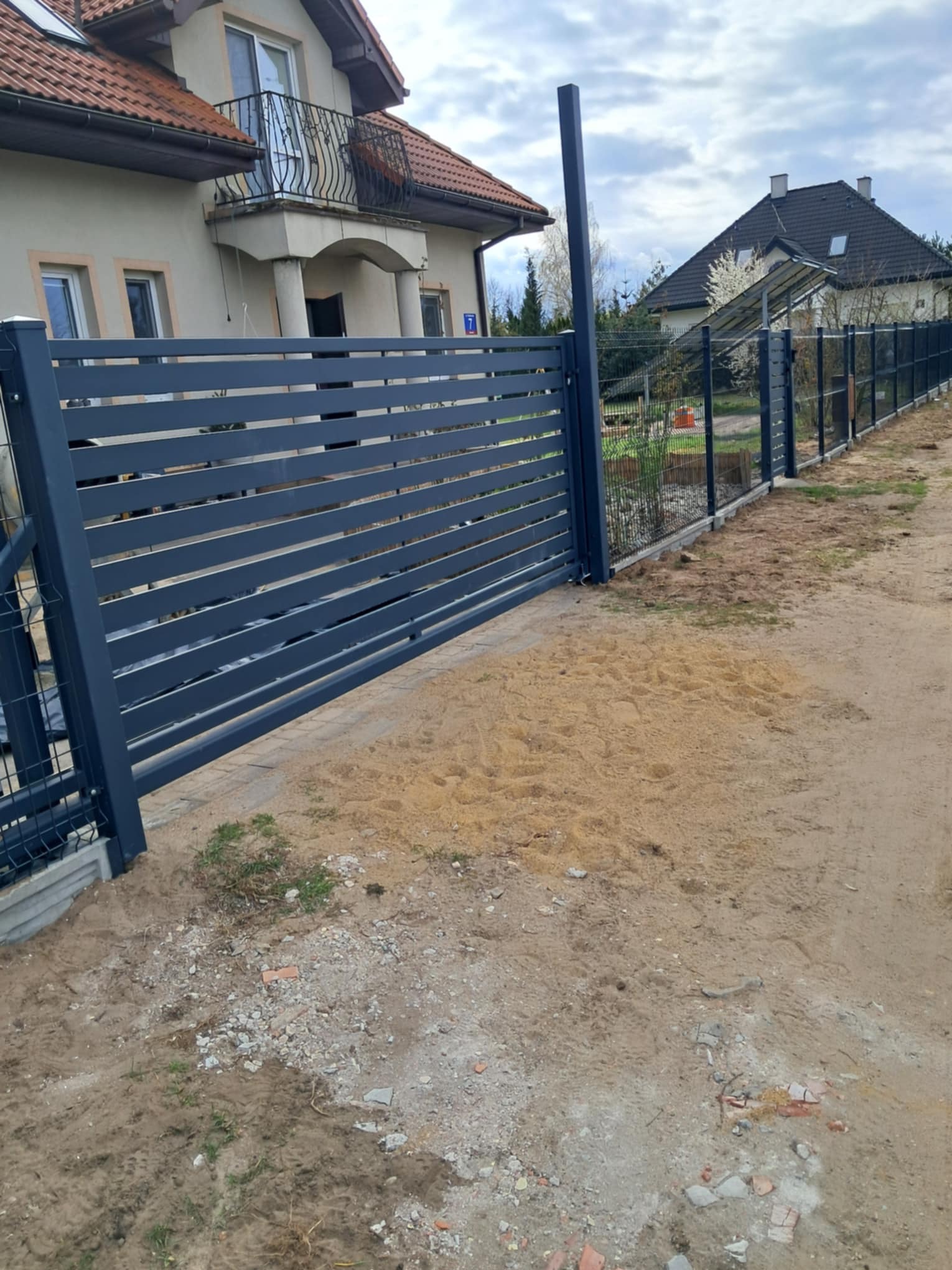 Nowoczesna, pozioma brama przesuwna z metalu w kolorze antracytowym. W tle widoczny dom jednorodzinny i fragment ogrodzenia panelowego.