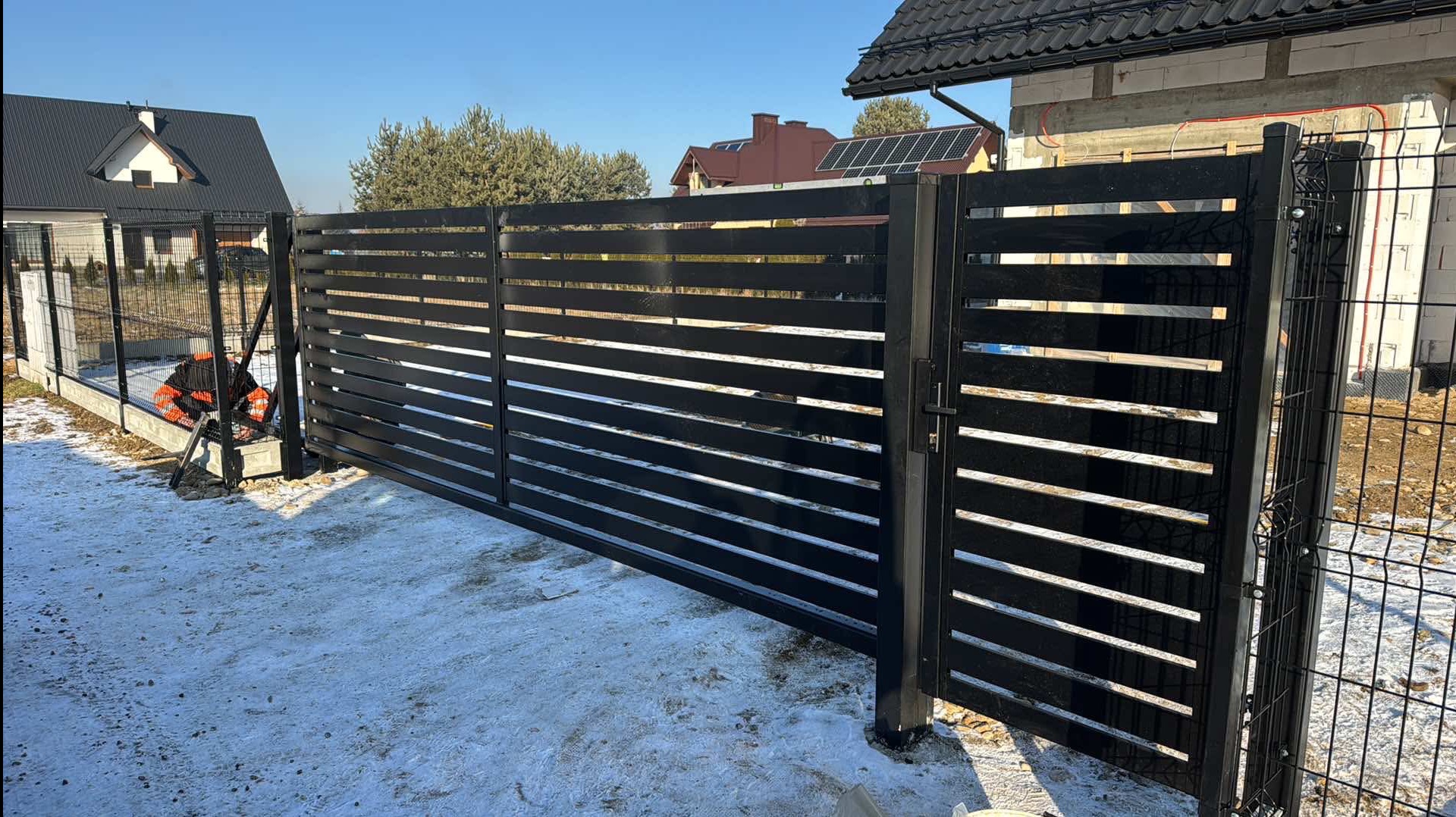 Nowoczesna, czarna brama przesuwna z poziomymi panelami na tle zimowego krajobrazu i domu z panelami słonecznymi na dachu. Widoczny fragment siatki ogrodzeniowej.