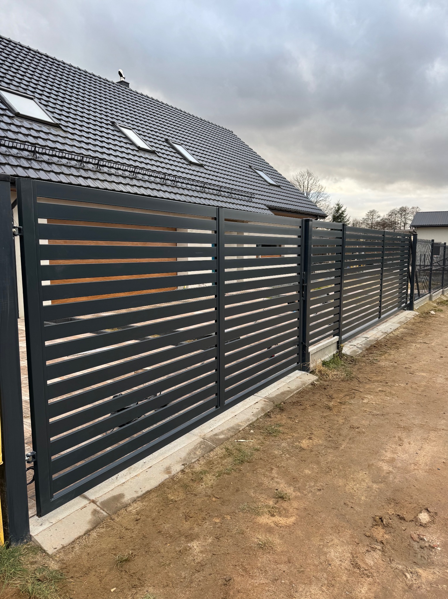 Nowoczesne, poziome, metalowe ogrodzenie panelowe w kolorze grafitowym, z bramą przesuwną, na tle domu z szarą dachówką i pochmurnego nieba.