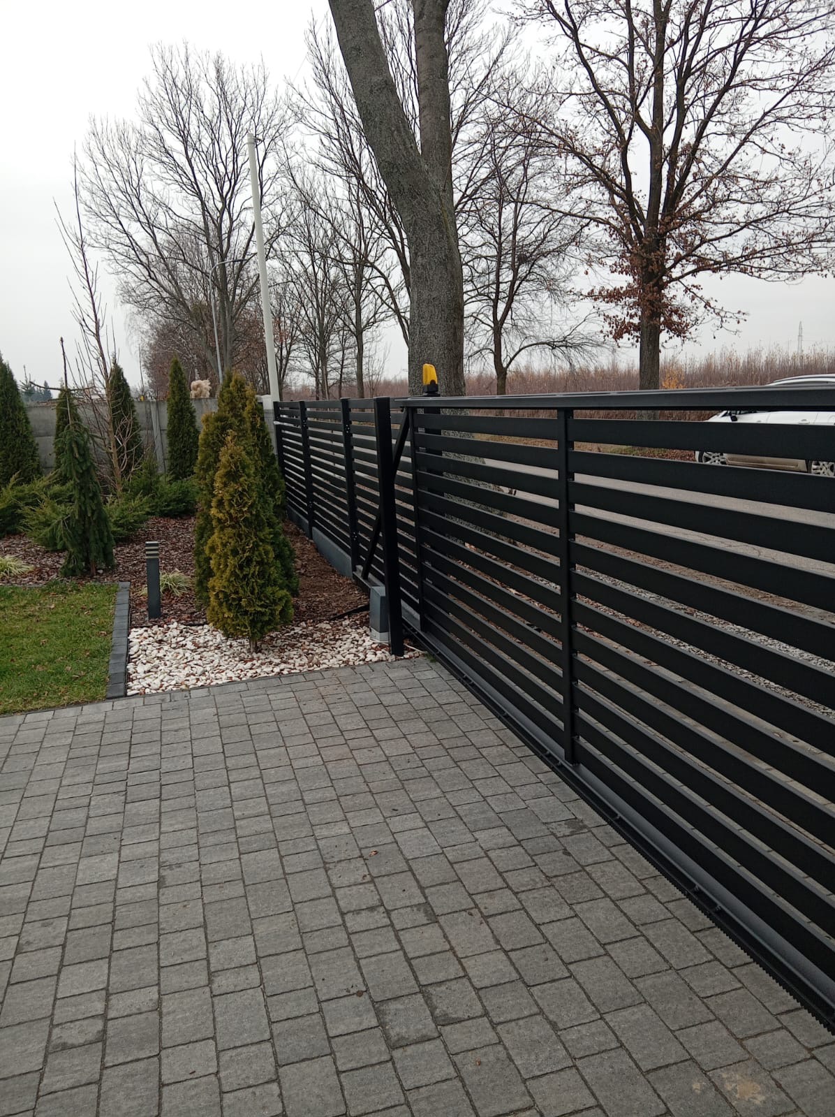Nowoczesne, poziome ogrodzenie panelowe w kolorze czarnym, z automatyczną bramą przesuwną. Widoczne kostki brukowe i ozdobne rośliny.