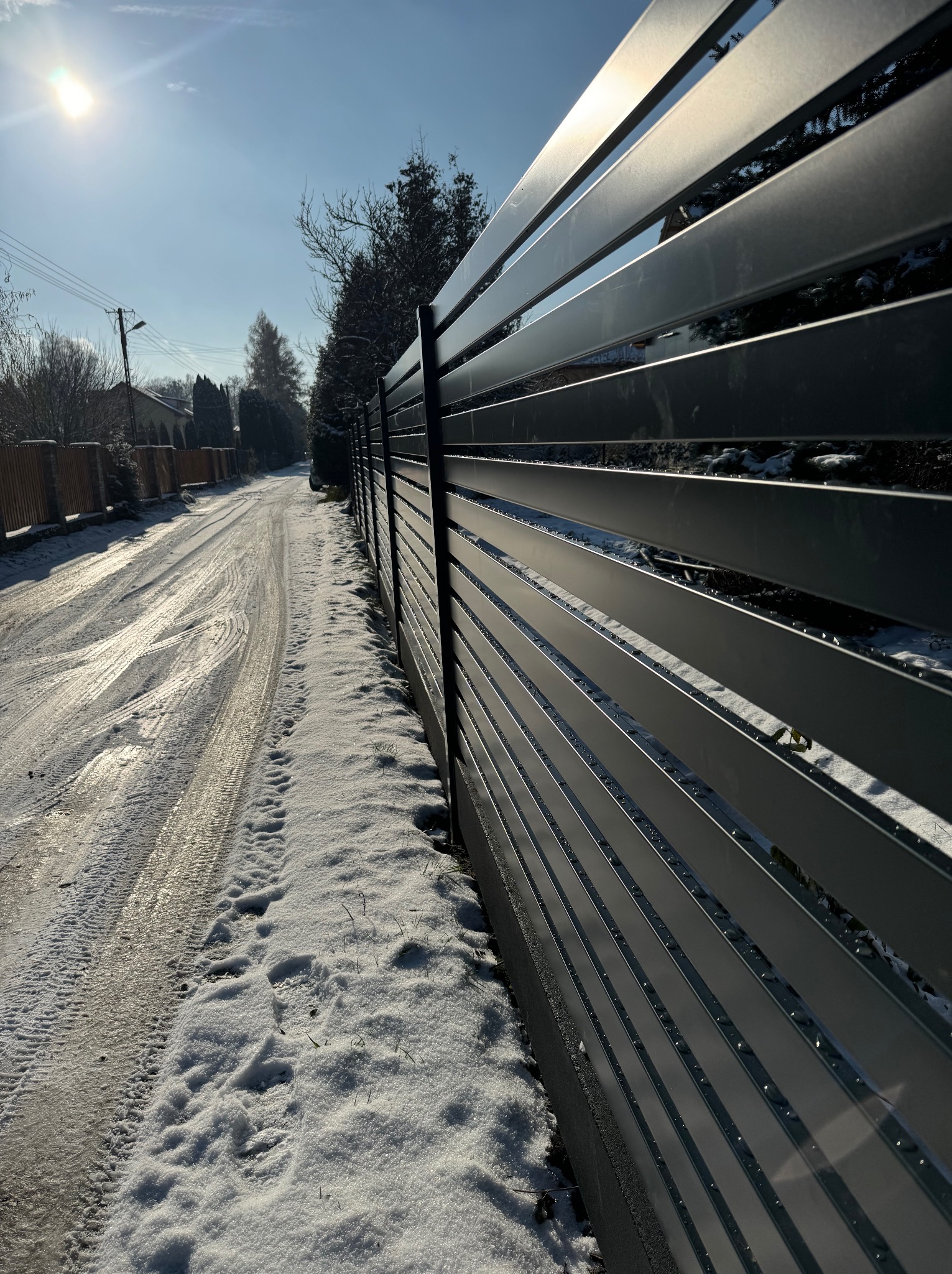 Nowoczesne, poziome przęsła metalowe ogrodzenia w zimowej scenerii. Śnieg na drodze i wzdłuż ogrodzenia. Słoneczny dzień.