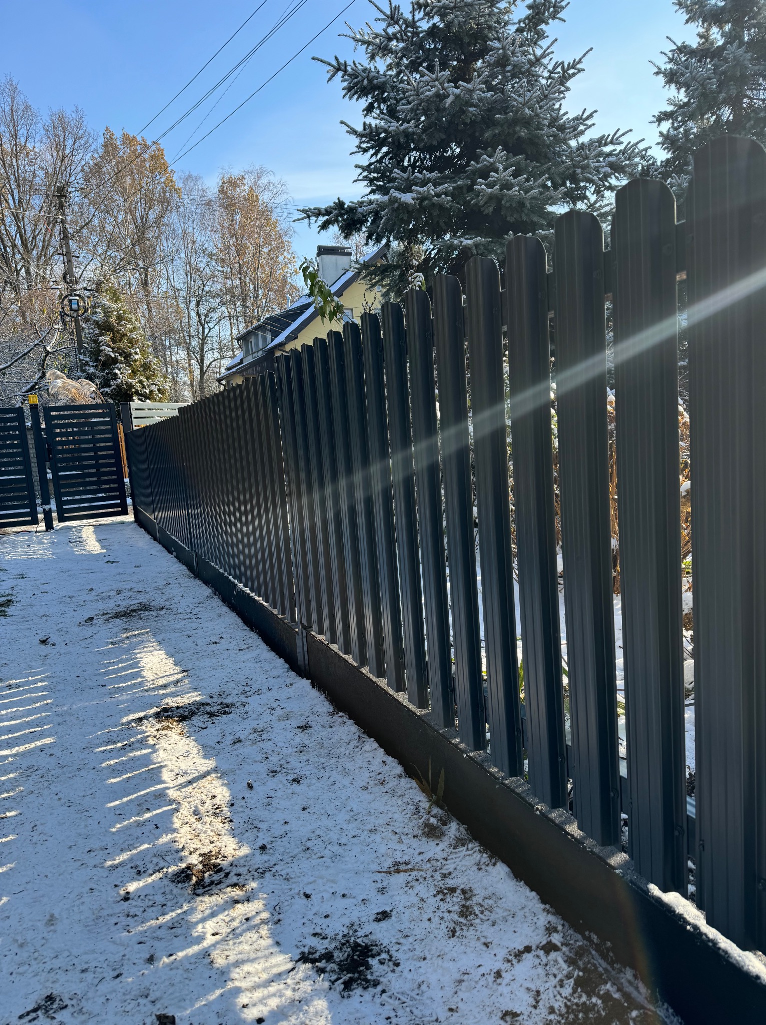 Nowoczesne, pionowe, metalowe ogrodzenie panelowe w kolorze grafitowym, otoczone śniegiem. Widoczny fragment furtki. Słoneczny dzień.