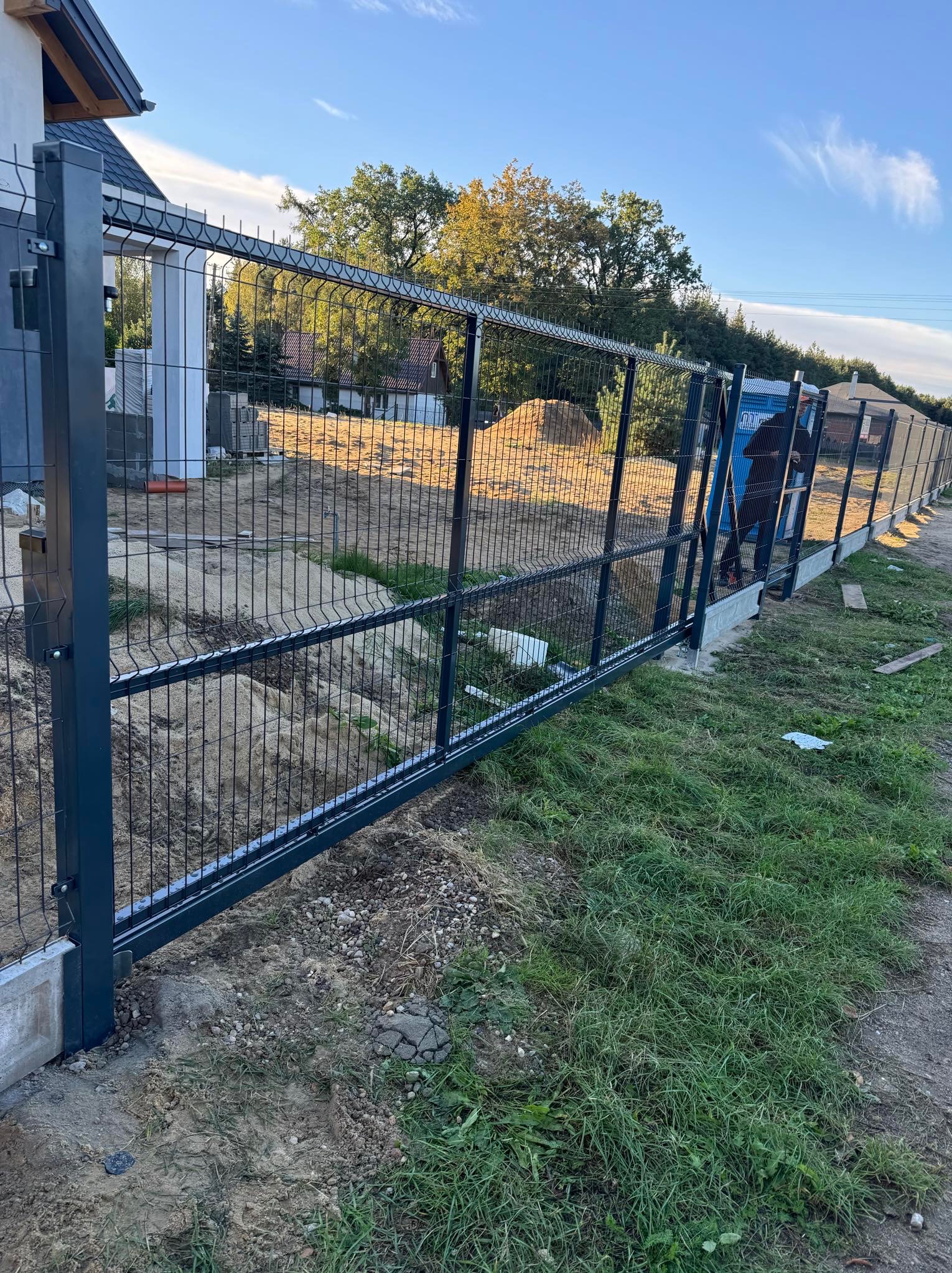 Nowoczesne ogrodzenie panelowe w kolorze grafitowym, z widoczną bramą przesuwną, zamontowane na betonowej podmurówce. W tle zielona trawa i drzewa.