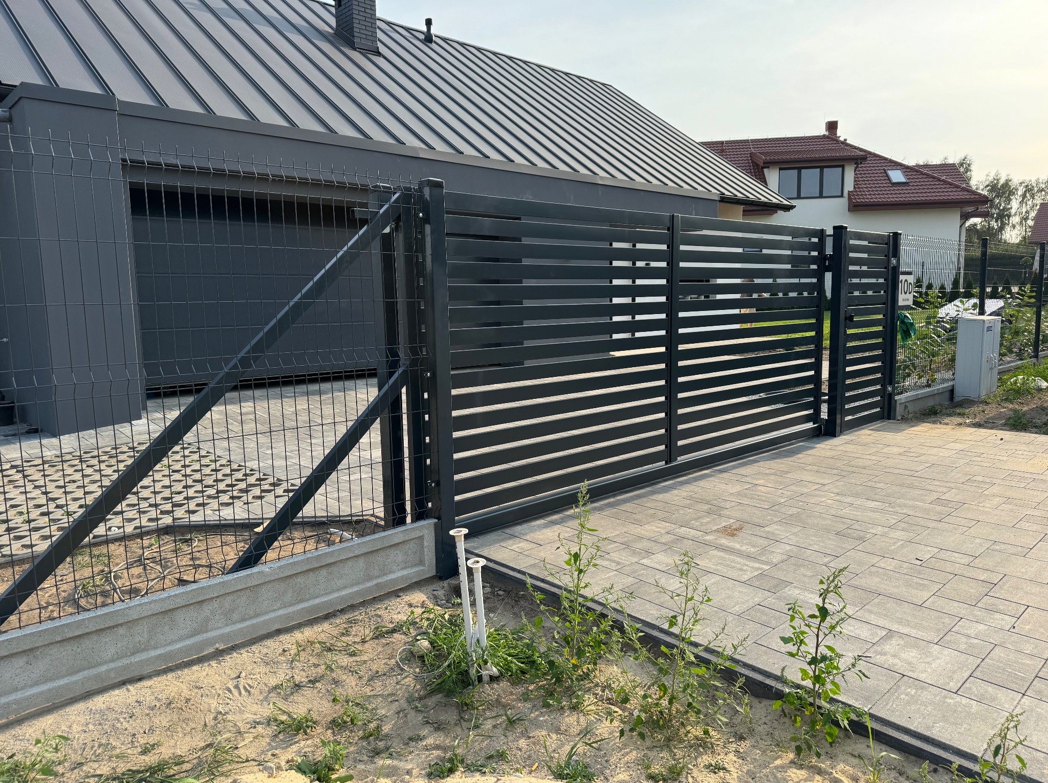 Nowoczesna, pozioma brama przesuwna z metalowymi panelami, w kolorze grafitowym, z widoczną siatką ogrodzeniową i kostką brukową na podjeździe.