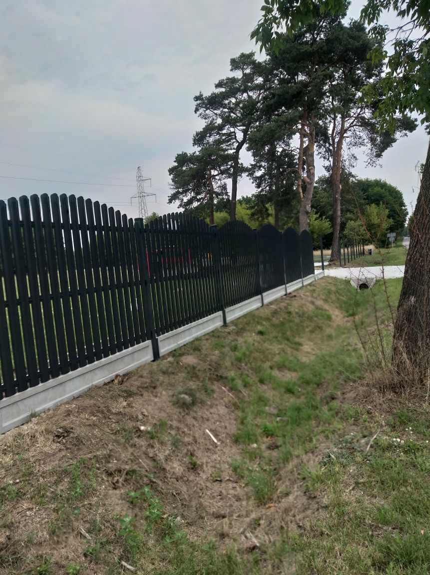 Nowoczesne ogrodzenie palisadowe w kolorze grafitowym, z podmurówką betonową, na tle zieleni i drzew. Widoczne słupy energetyczne w oddali.