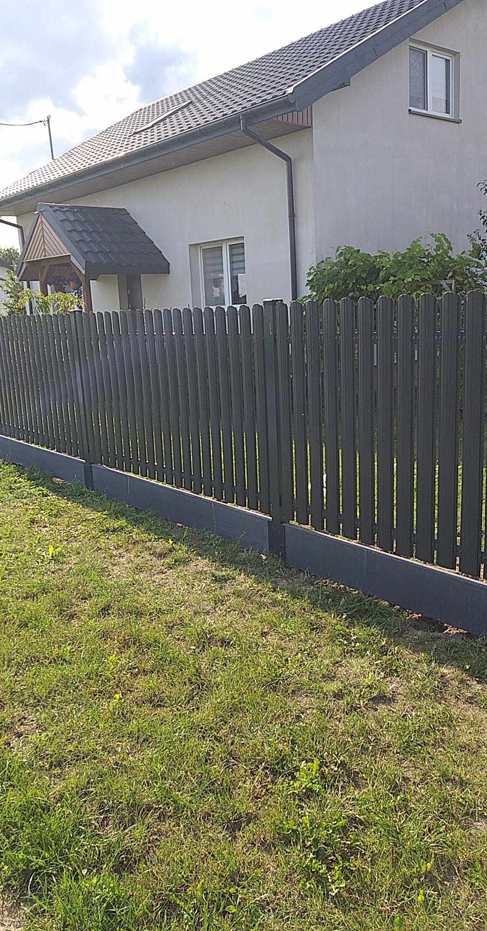 Nowoczesne ogrodzenie metalowe w kolorze grafitowym, z podmurówką, na tle domu jednorodzinnego i trawnika w słoneczny dzień. Widok z poziomu gruntu.