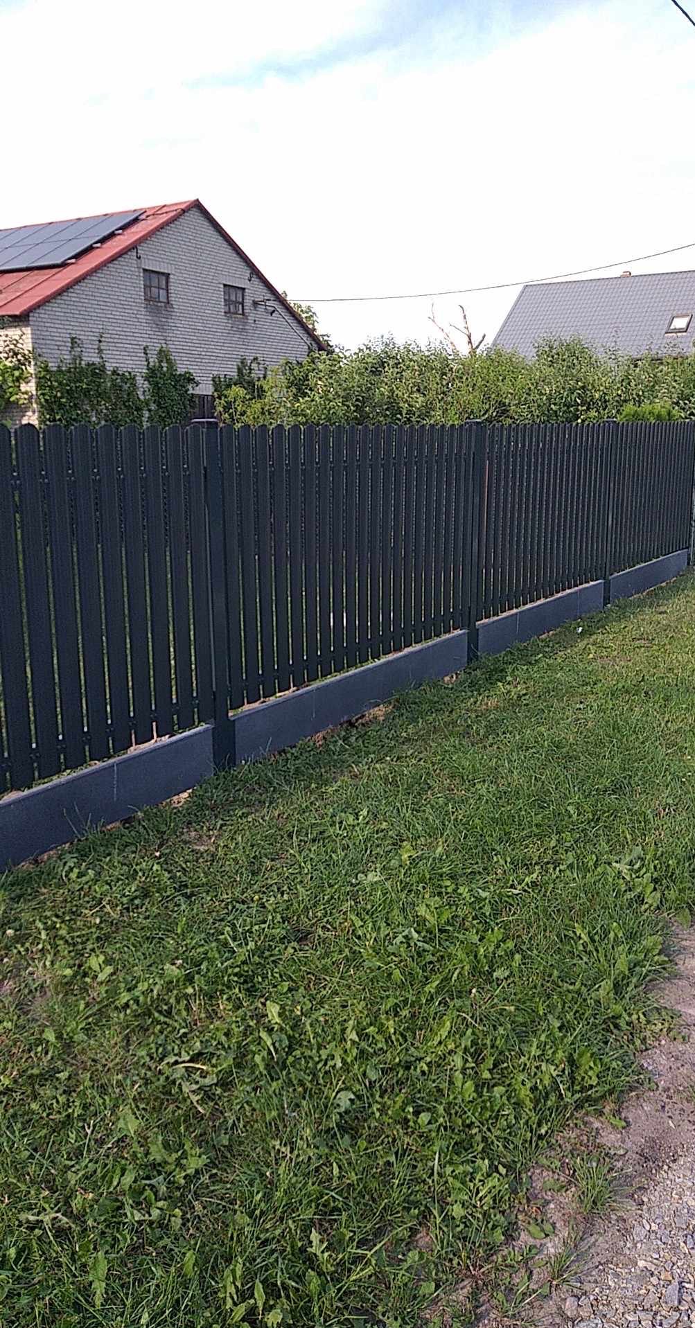 Ciemny, pionowy płot panelowy z podmurówką na tle zielonej trawy i budynków z dachami; widok z poziomu gruntu.