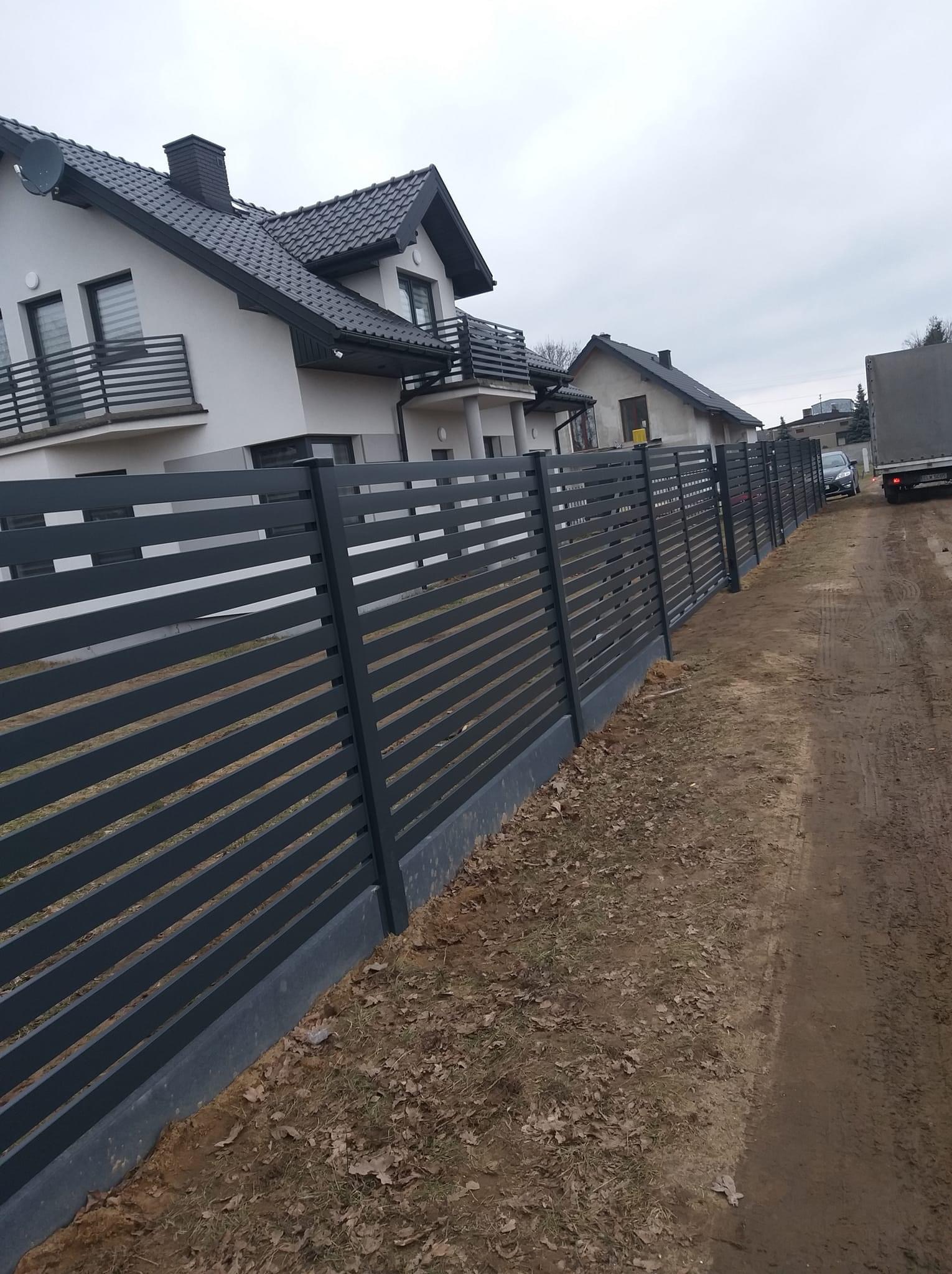 Nowoczesne, poziome ogrodzenie panelowe w antracytowym kolorze, oddzielające posesję z nowym domem. Widoczny fragment drogi i zaparkowany samochód.
