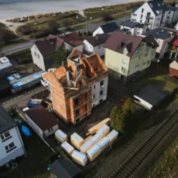 Budowa domu z czerwonej cegły na działce z widokiem z lotu ptaka. W tle morze i zabudowa miejska. Częściowo otynkowane ściany i widoczne pustaki.