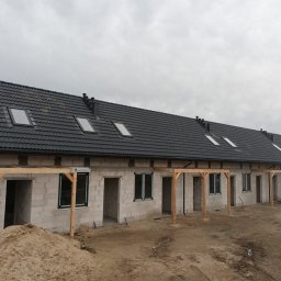 BUD ROB - Szeregówka w stanie surowym otwartym, z oknami, dachem z lukarnami i drewnianą konstrukcją werandy. Na pierwszym planie hałda piasku.