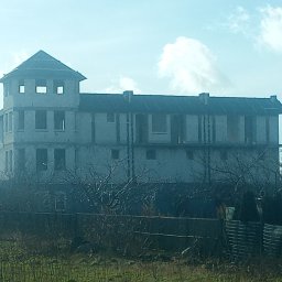 BUD ROB - Niedokończony, trzypiętrowy budynek z wieżą w stanie surowym, widoczny zza ogrodzenia i roślinności. Brak okien, surowy beton. Błękitne niebo w tle.