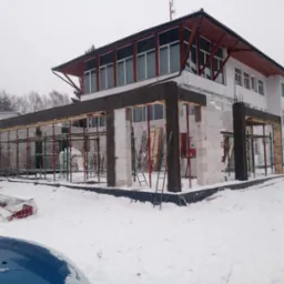 Budowa domu w zimowej scenerii: wznoszone ściany z białych bloczków, widoczne rusztowania i szalunki, tło stanowi biały budynek i zasypane śniegiem otoczenie.