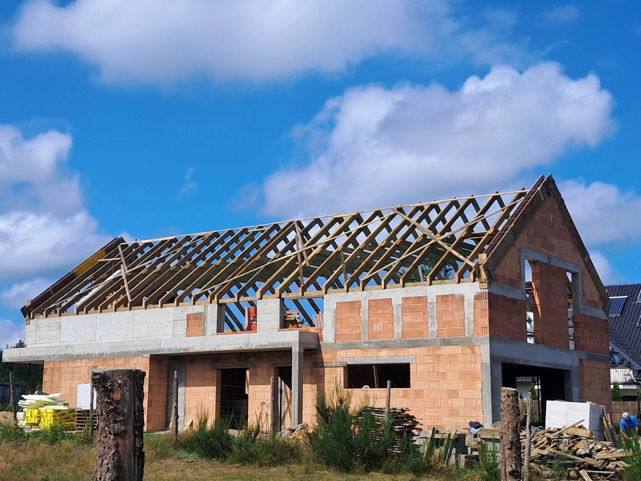 Surowy stan domu z czerwonej cegły, z drewnianą konstrukcją dachu i widocznymi elementami betonowymi, na tle błękitnego nieba z chmurami.