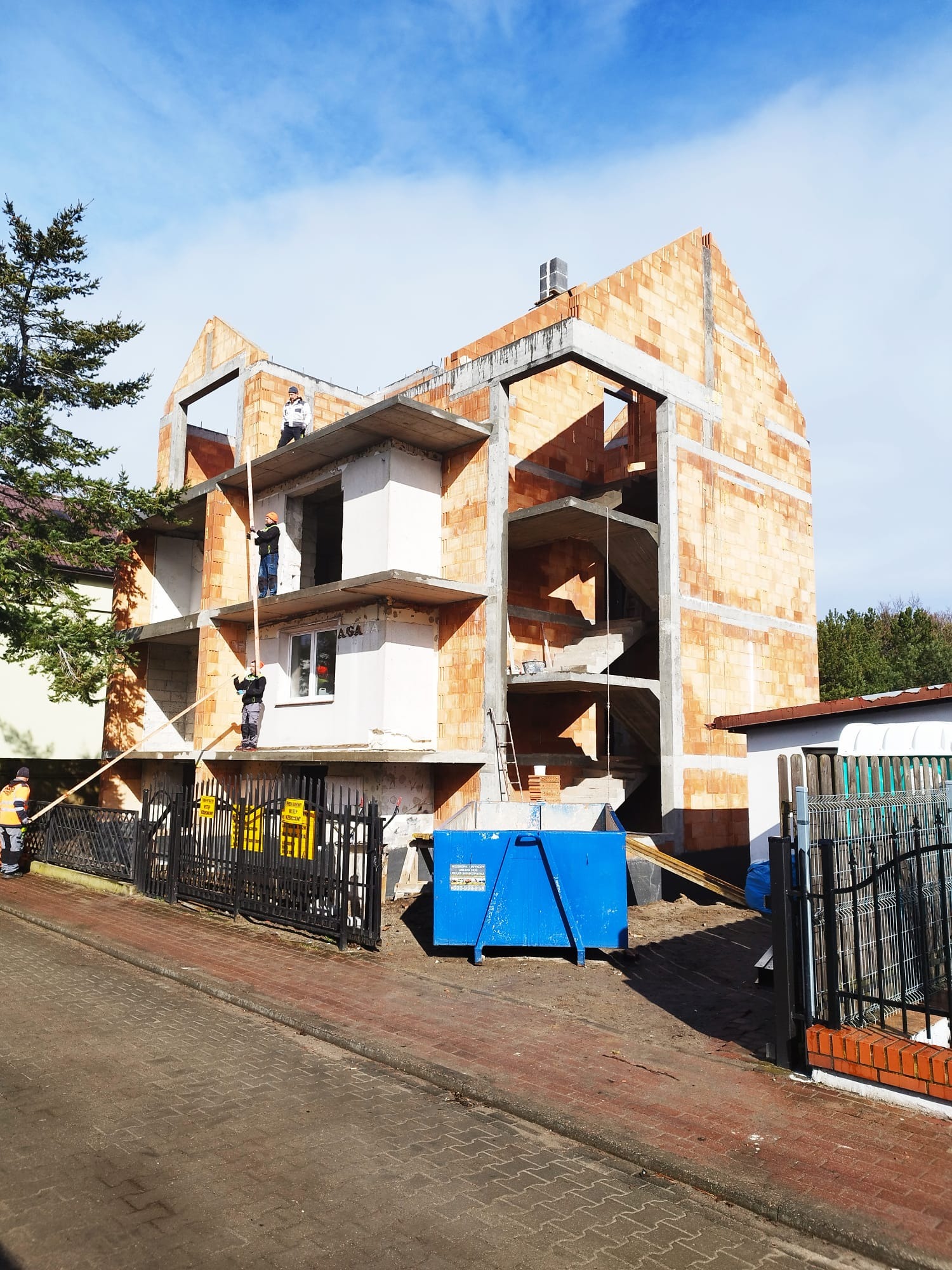 Wznoszenie trzypiętrowego domu z czerwonej cegły. Widoczni robotnicy na różnych poziomach konstrukcji, błękitny kontener na gruz, metalowe ogrodzenie i fragment chodnika.